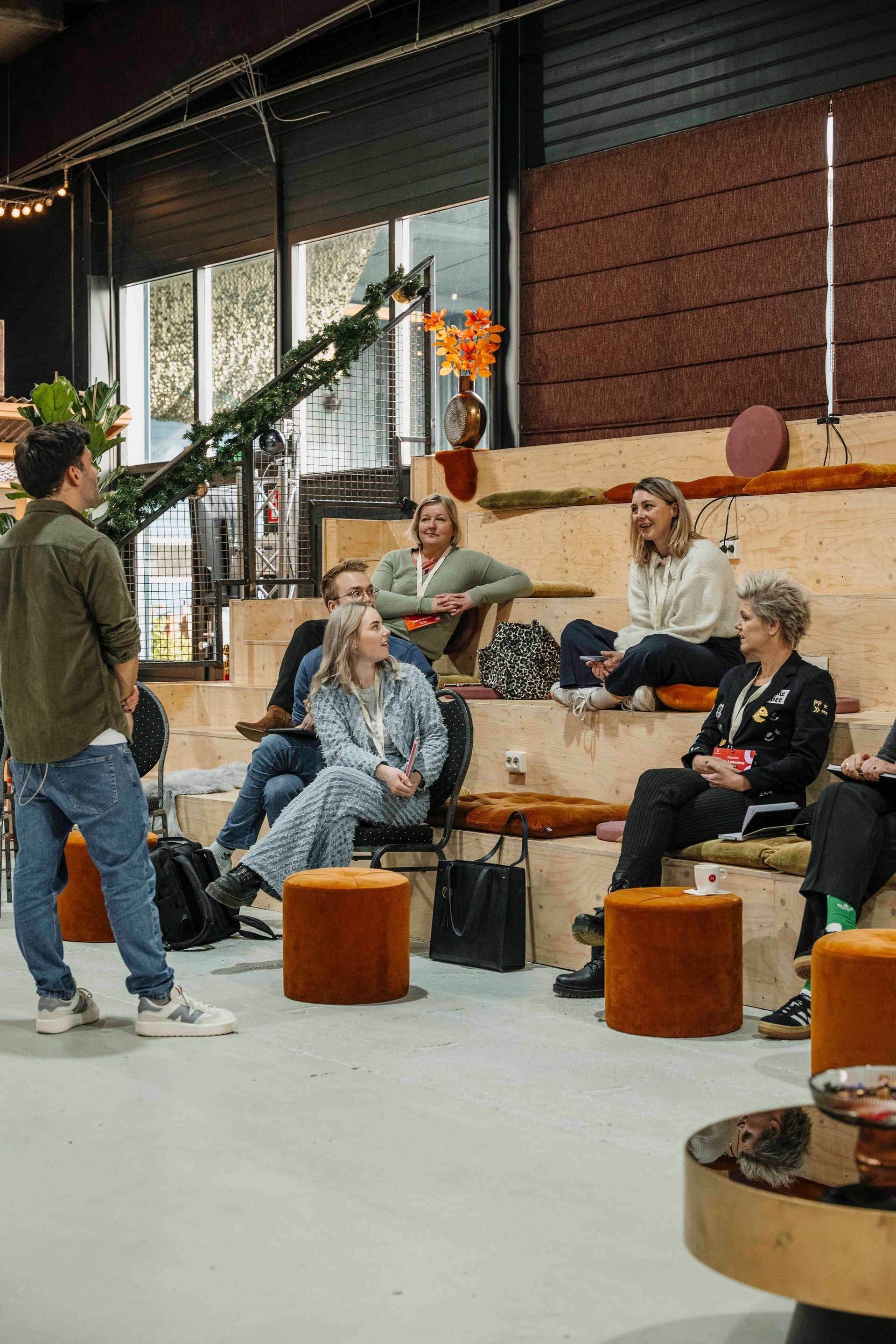 Een spreker spreekt een groep mensen toe die op een houten trap zitten in een informele, helder verlichte binnenruimte.