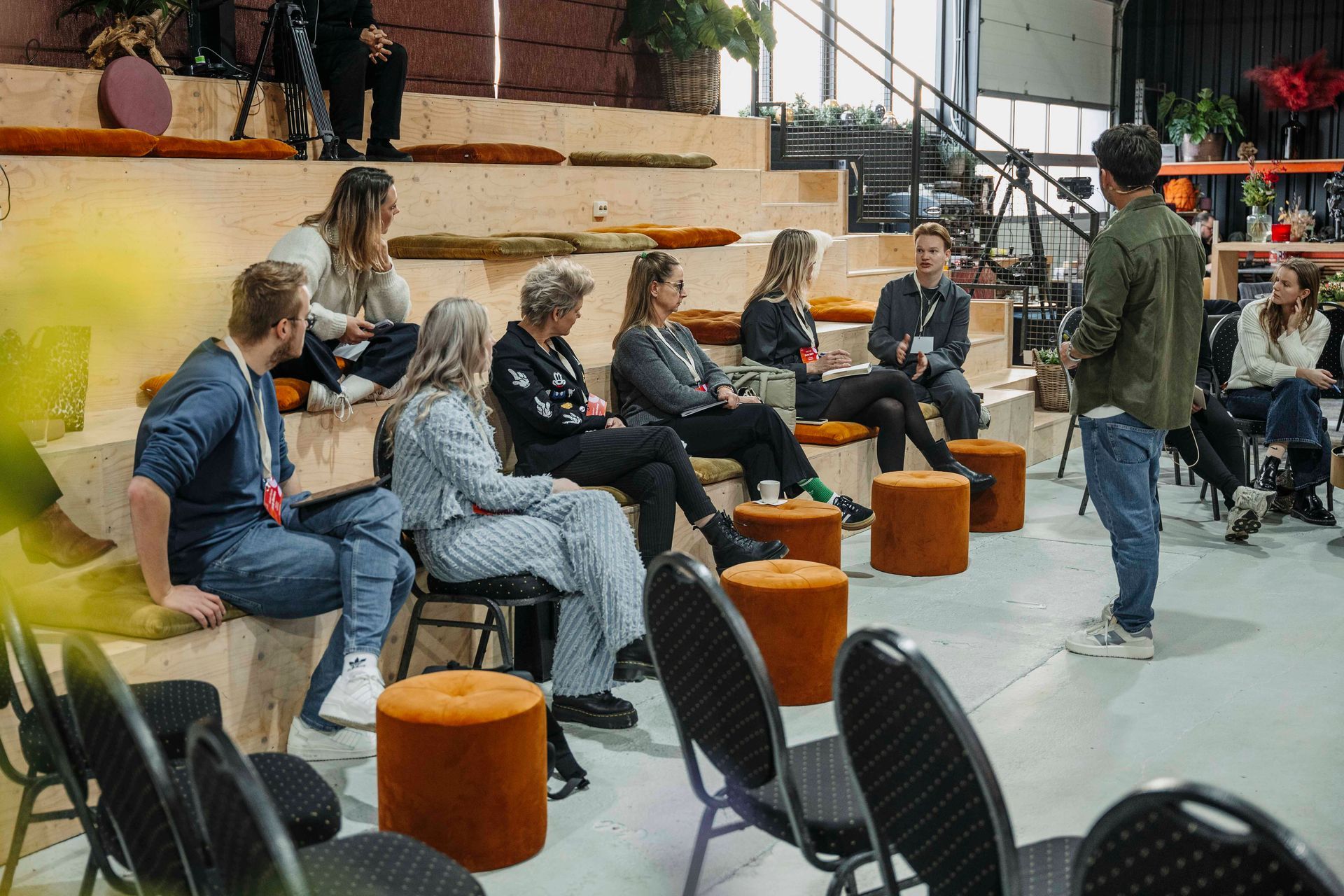Een groep mensen zit op houten tribunezitjes in een studio en luistert naar iemand die staat en een presentatie geeft.