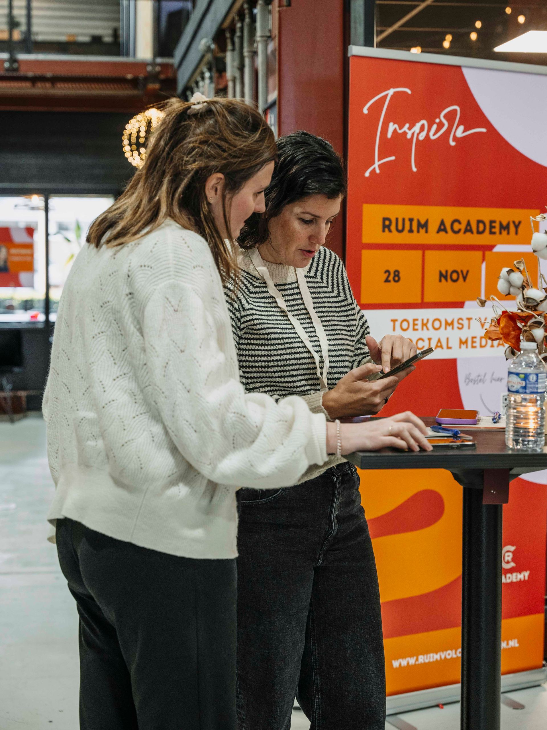 Twee mensen kijken samen naar een smartphone naast een evenementbanner van RUIM Academy in een binnenruimte.