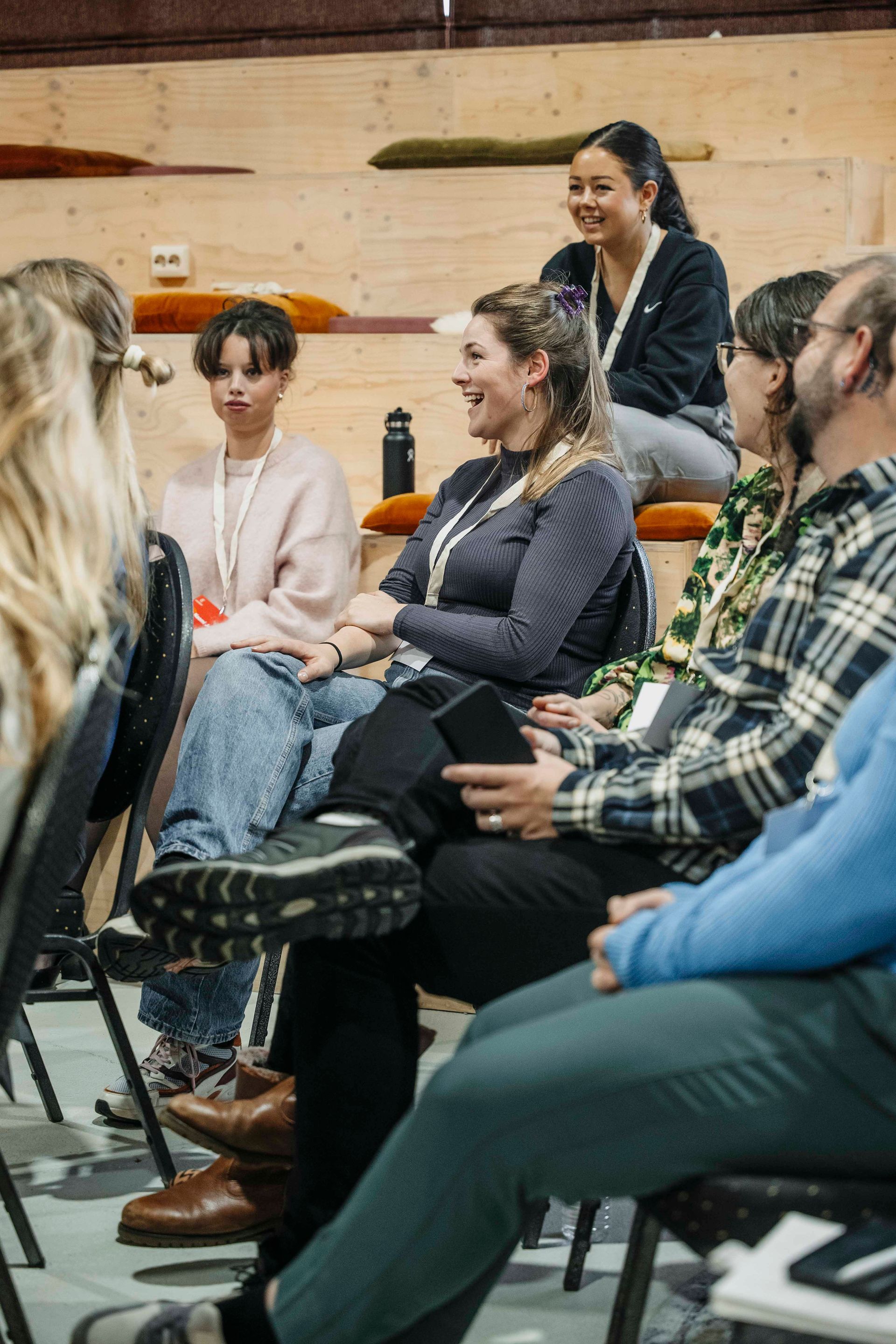 Een groep mensen zit in een binnenruimte met houten tribunezitplaatsen, glimlachend en in gesprek.