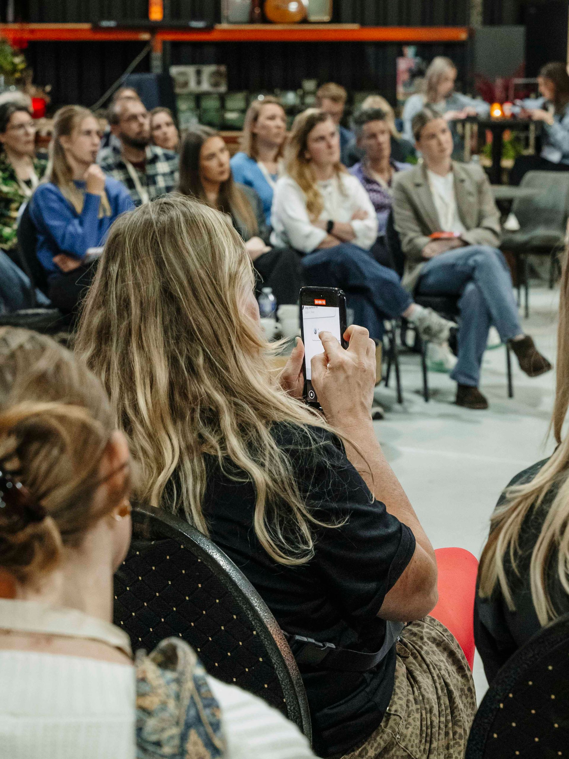 Tijdens een presentatie voor een zittend publiek in een zaal in de stijl van een loods, gebruikt een persoon op de voorgrond een smartphone.