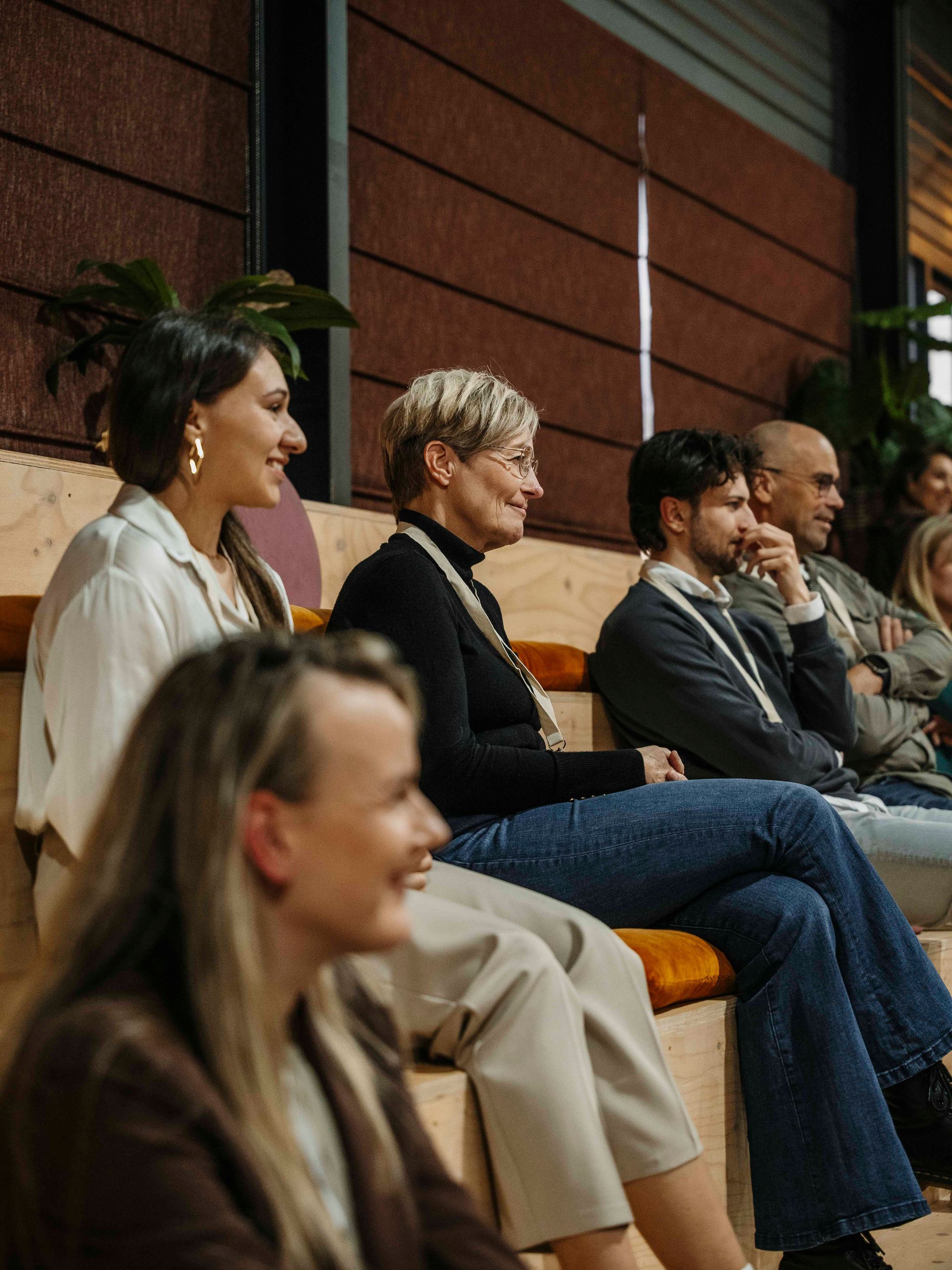 Een diverse groep mensen zit op een houten tribune en luistert en kijkt naar een evenement in een moderne binnenruimte.
