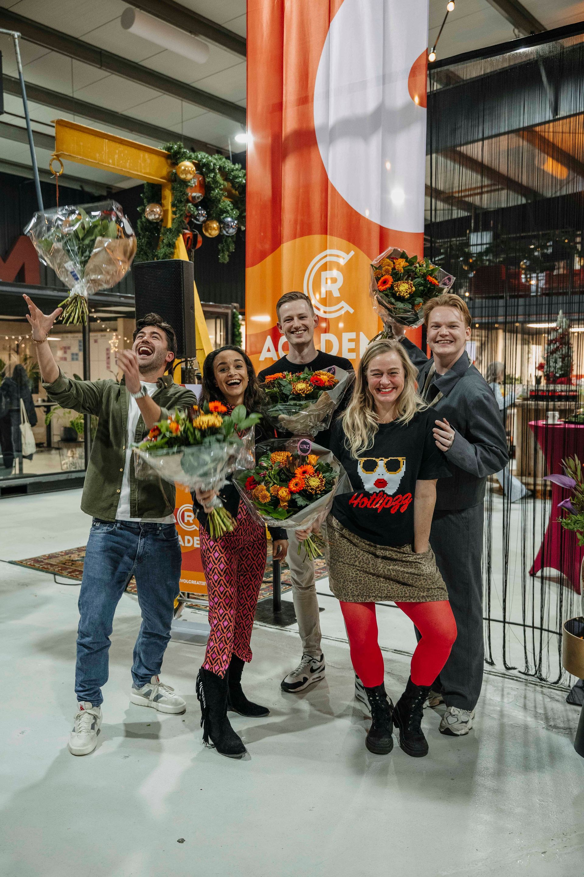 Vijf mensen met boeketten in hun handen vieren feest voor een groot oranje spandoek in een felverlichte, industriële ruimte.