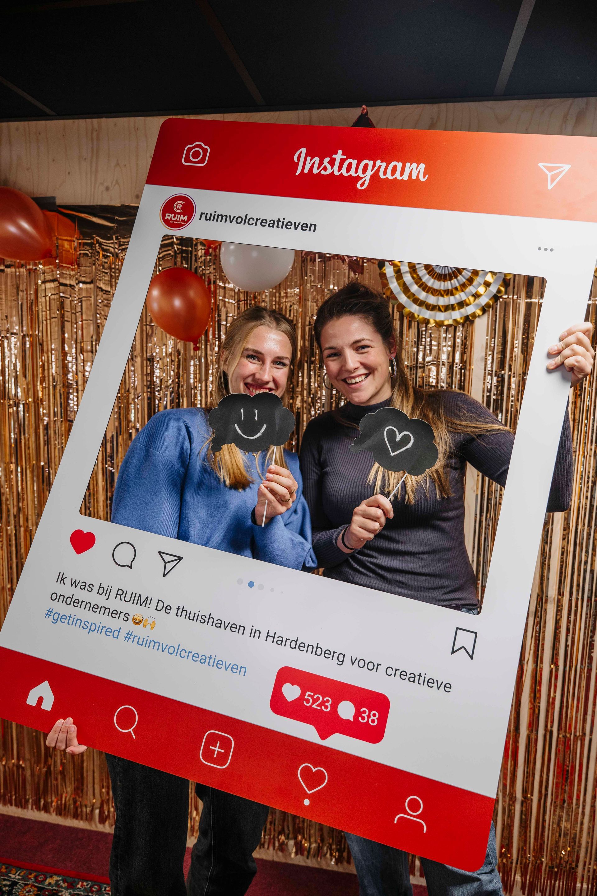 Twee mensen houden een groot kartonnen frame met Instagram-thema vast voor een achtergrond van gouden slingers.
