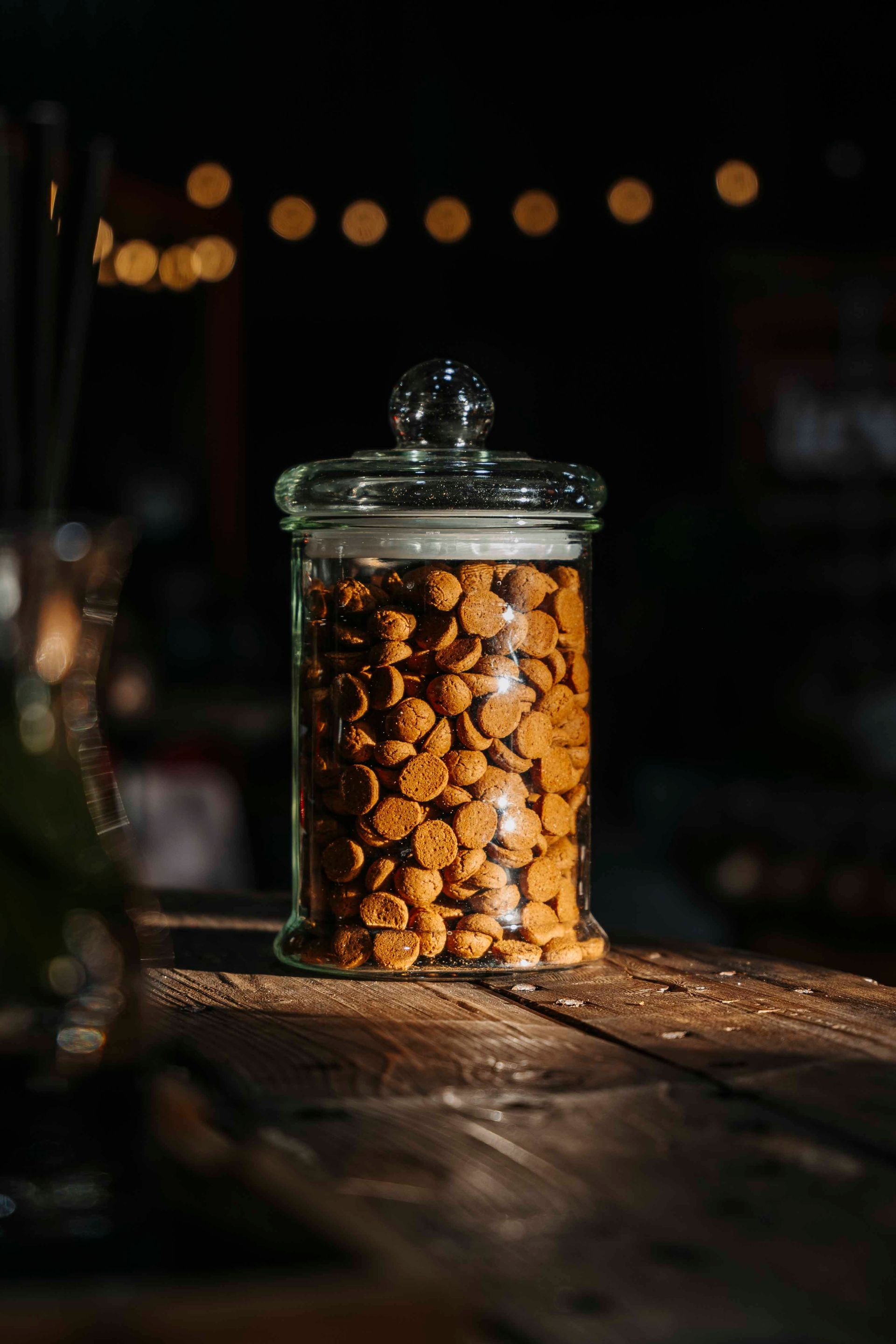 Een glazen pot gevuld met kleine, goudbruine ronde crackers staat op een houten tafel, verlicht door warm bokeh-achtergrondlicht.
