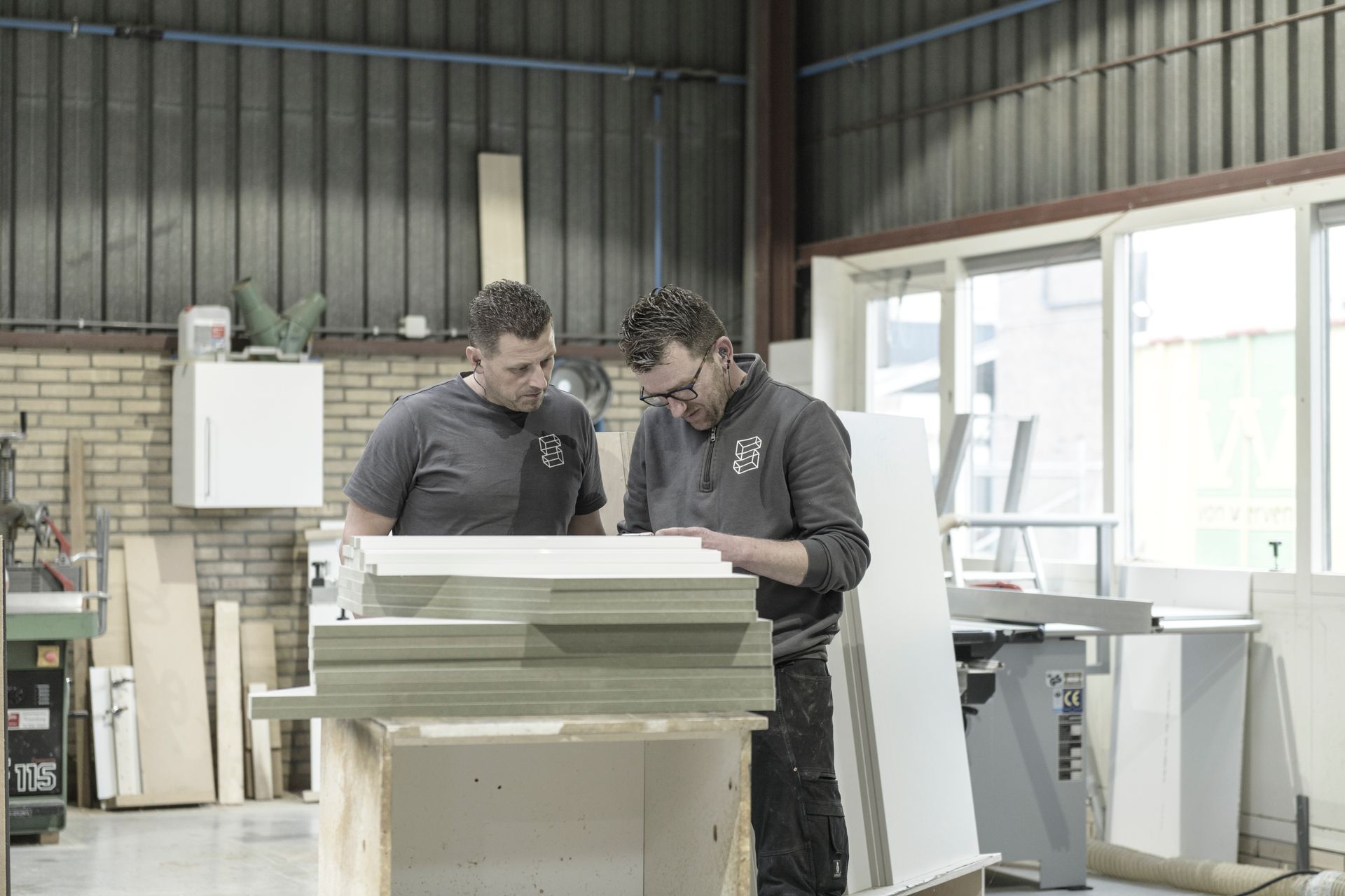 Twee mannen staan in een werkplaats naast elkaar en kijken naar een stuk hout.