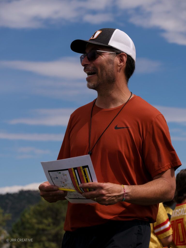 Person in an orange shirt and cap holding a paper, standing outdoors under a blue sky.