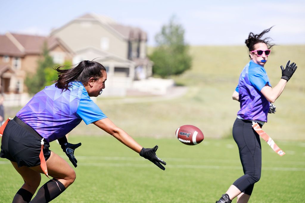 Two football players in purple jerseys chase a ball on a grassy field near houses.