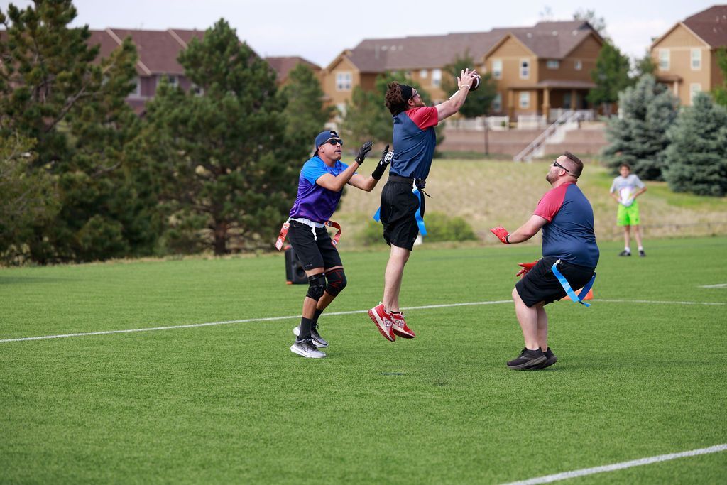 Three players jump for a catch on a grassy field with houses in the background.