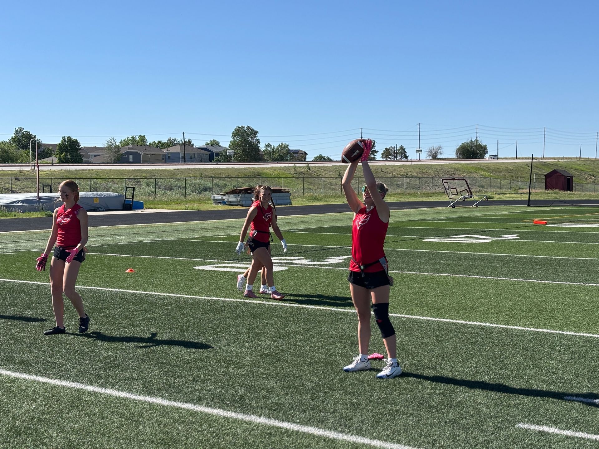 Girls playing flag football on a sunny field, with one player raising the ball overhead.