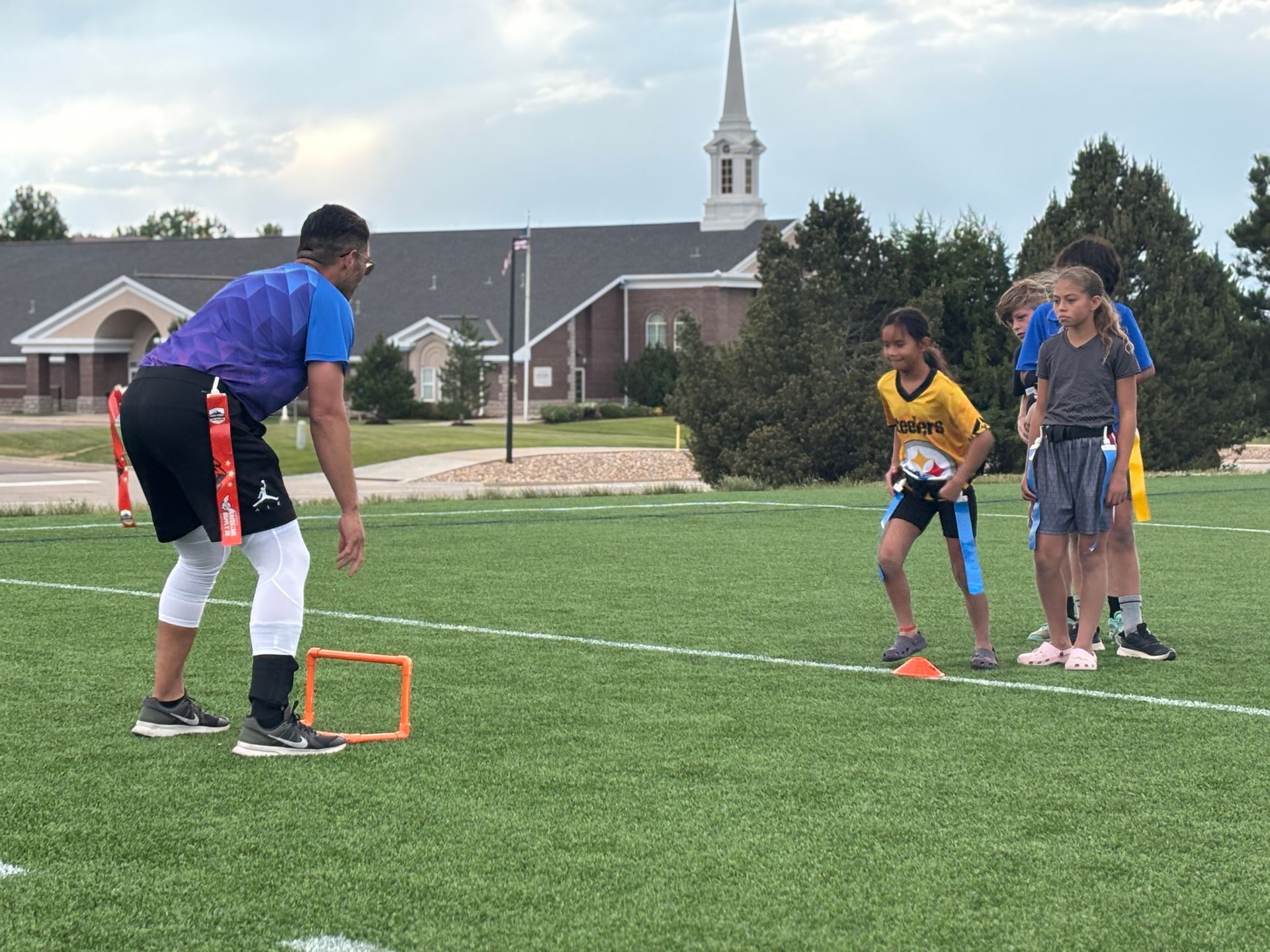 Coach sets up a drill as three kids watch on a soccer field, with a church steeple in the background