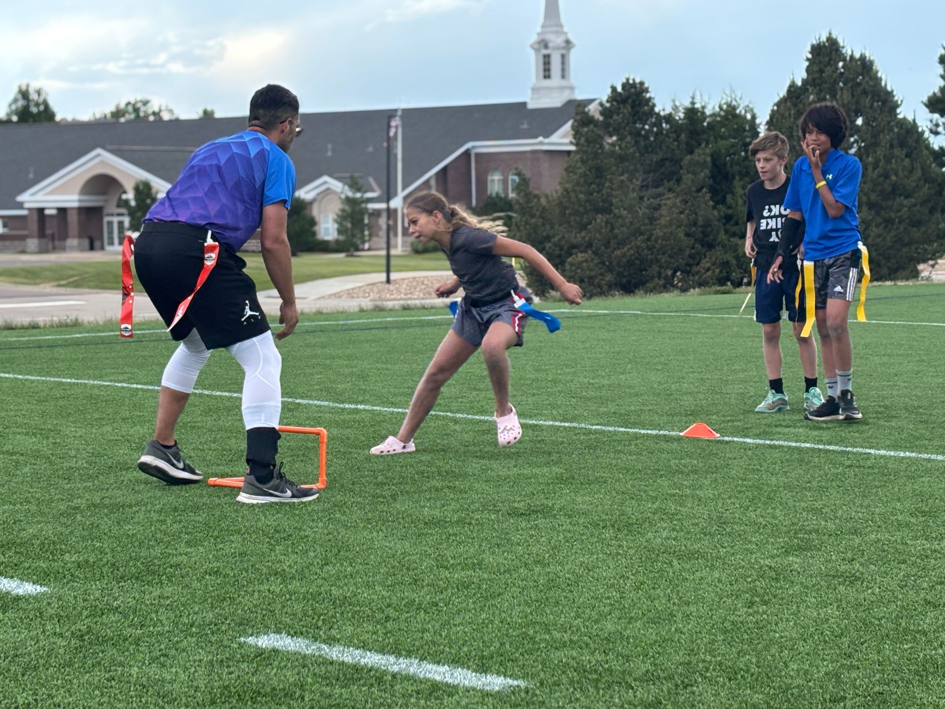 Children playing flag football on a field, with cones and a coach in blue guiding a drill.