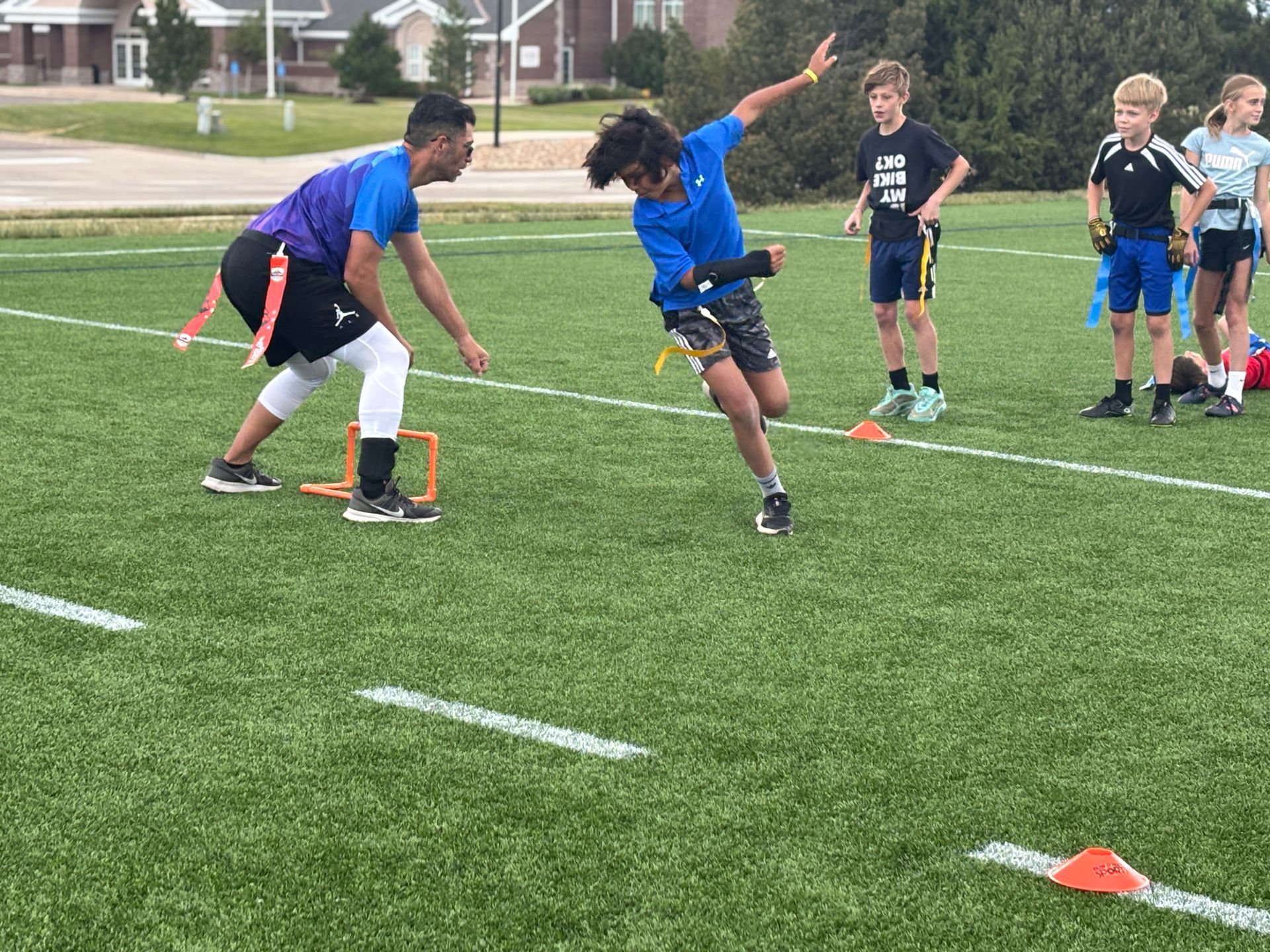 Children doing a relay drill on a grassy sports field with orange cones and two runners in blue shirts passing a baton