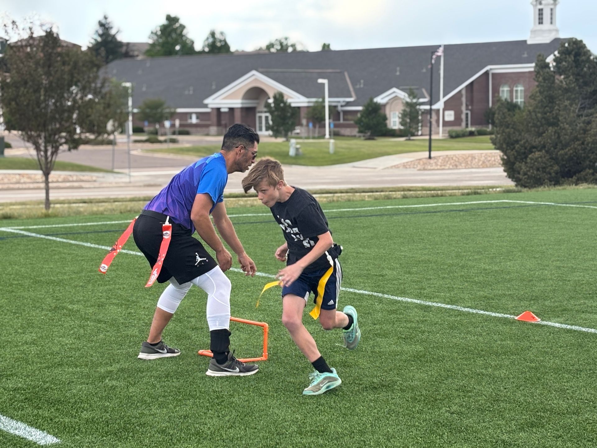 Two boys playing flag football on a grassy field with orange cones and houses in the background