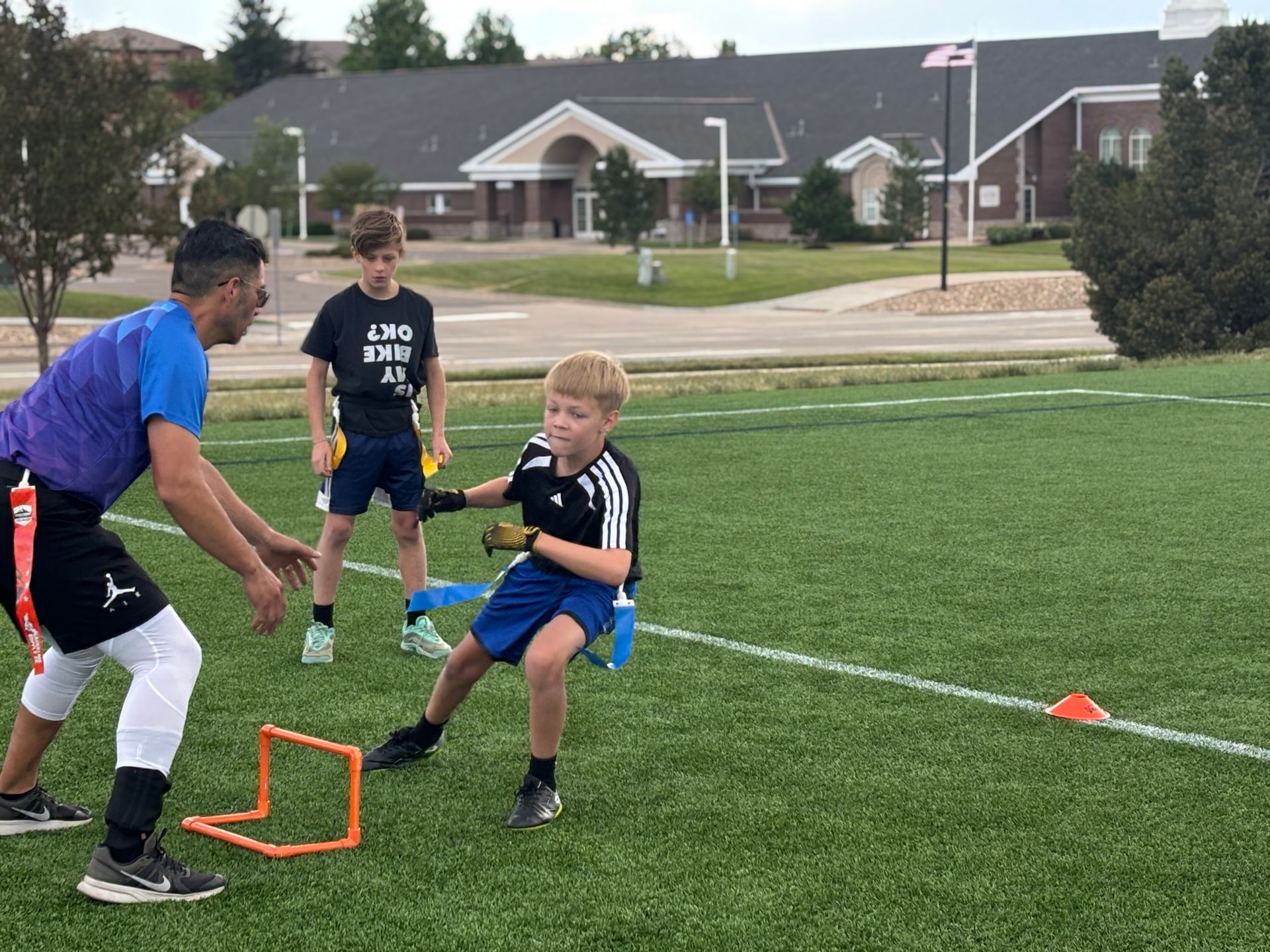 Kids practice football drills on a grassy field with a coach, orange hurdle, and cones.