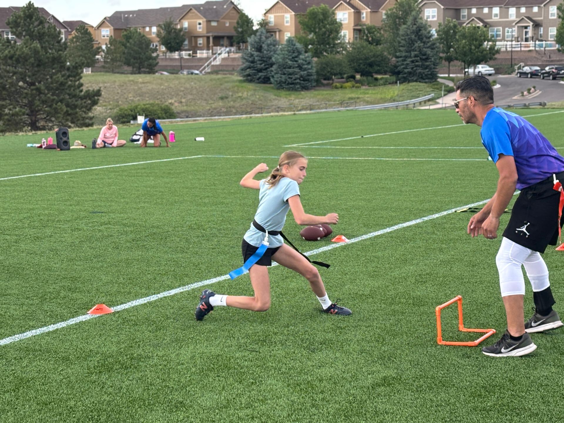 Girl running with a football during a youth practice drill on a grassy field, coach nearby and orange hurdles set up