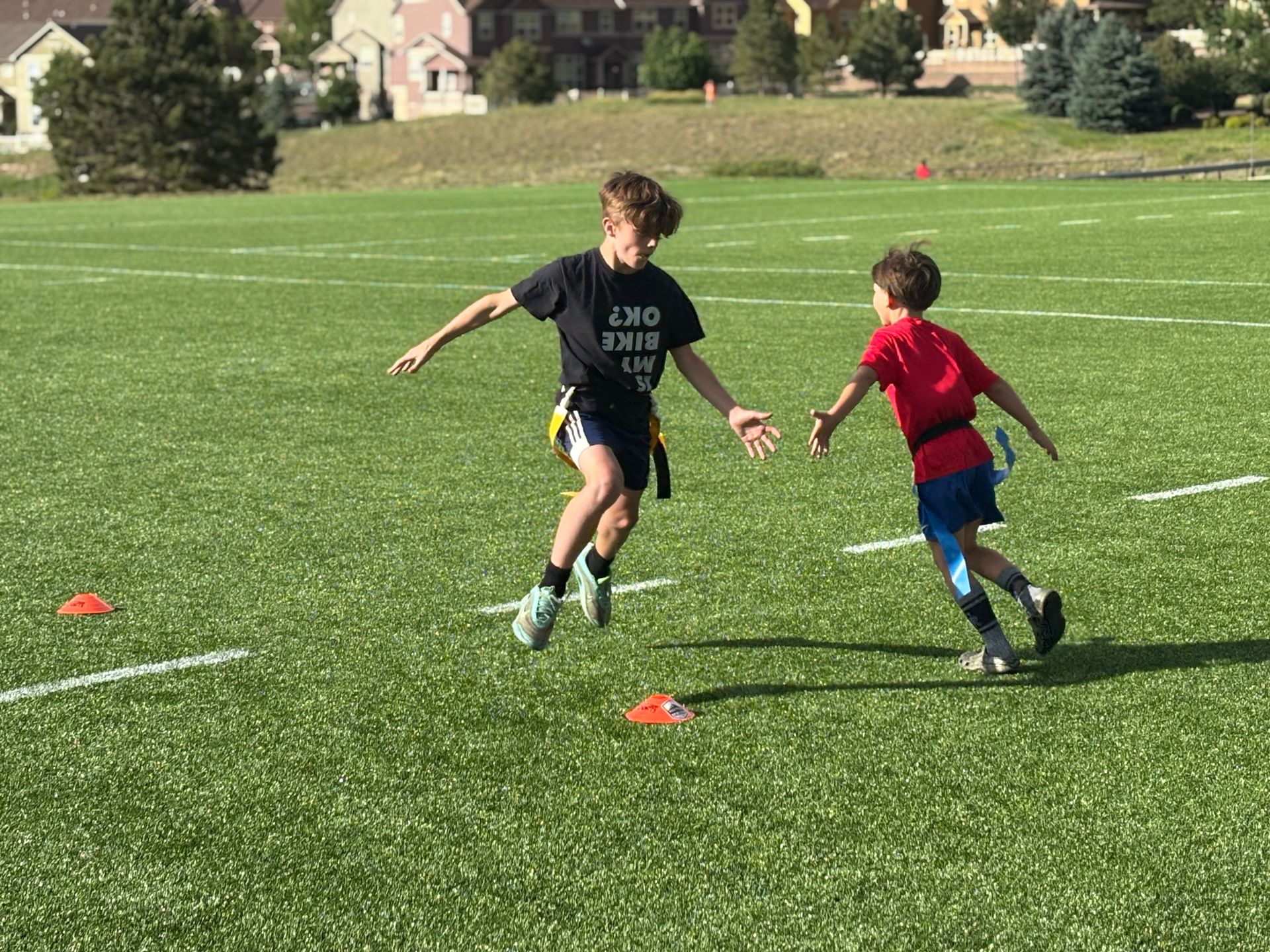 Two children in sports clothes running on a grassy field with orange cones