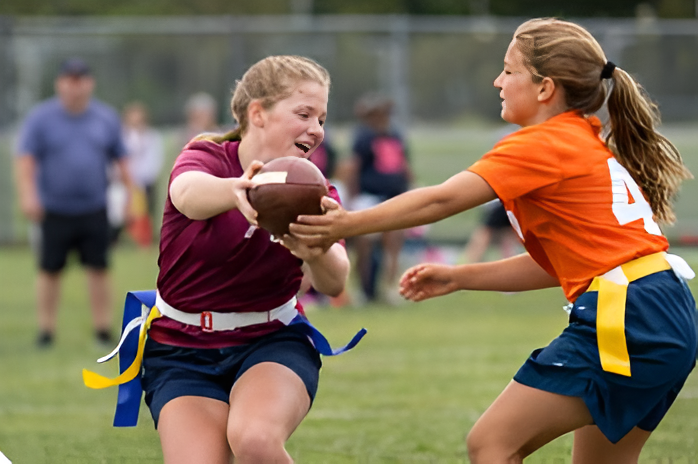 Two flag football players in maroon and orange reach for the ball on a grassy field.