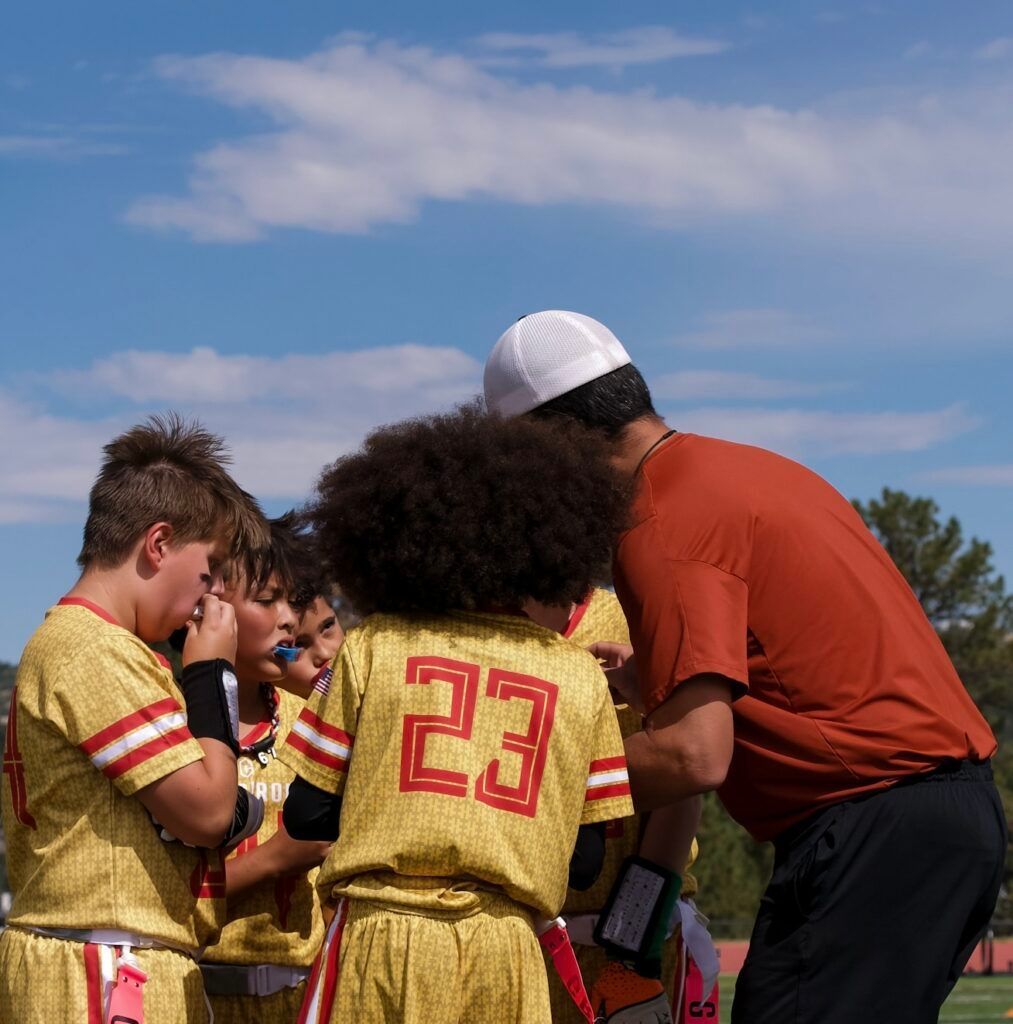 Youth soccer coach in orange shirt huddles with players in gold jerseys on a sunny field