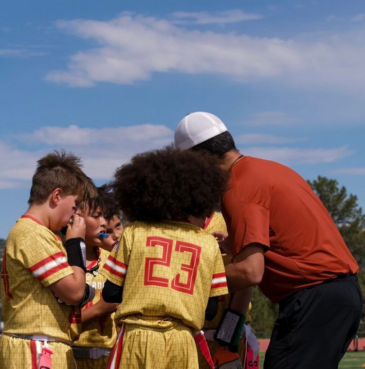 Coach talking to young football players on a field under a blue sky
