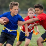 Kids playing flag football on a field, one in blue running with the ball as another in red reaches for him