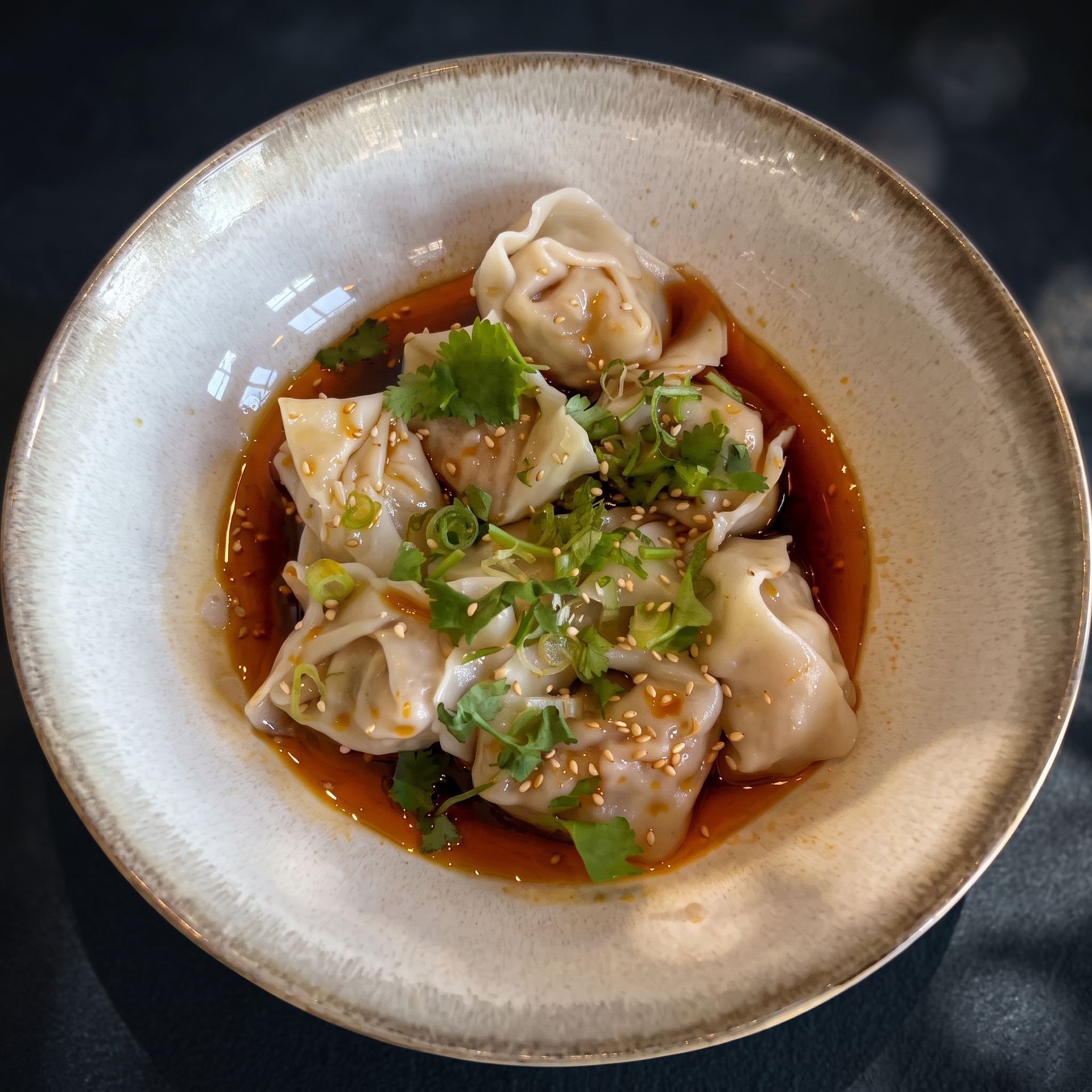 Wonton dumplings in a bowl of soy-based broth, garnished with cilantro and sesame seeds.