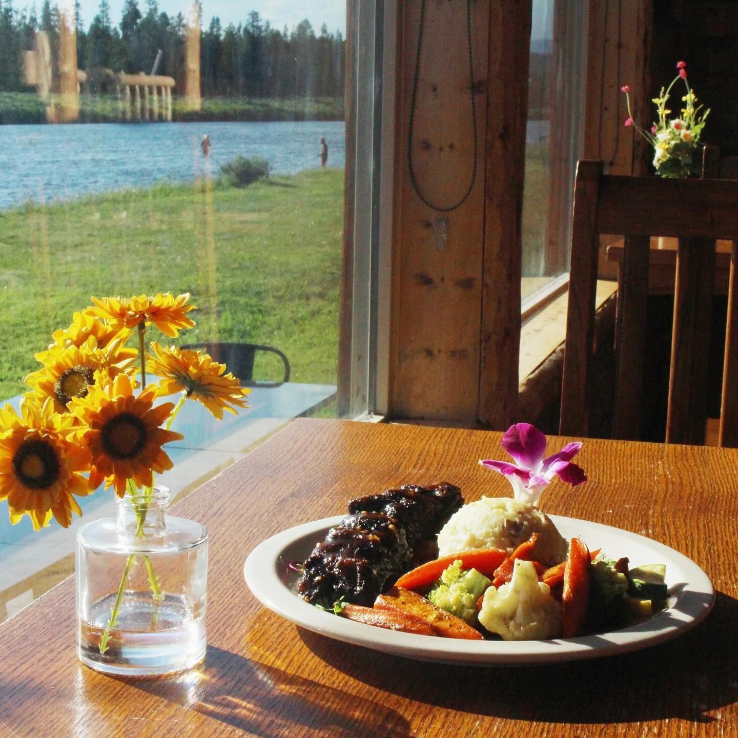 Meal of meat, mashed potatoes, and vegetables on a plate, beside sunflowers, with a lake view from a window.