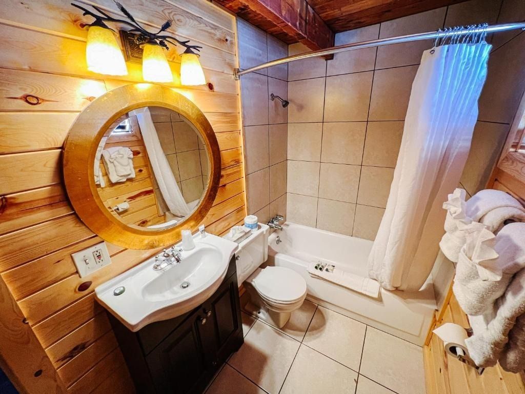 Bathroom with wooden walls, sink, toilet, tub/shower, round mirror, and towels.