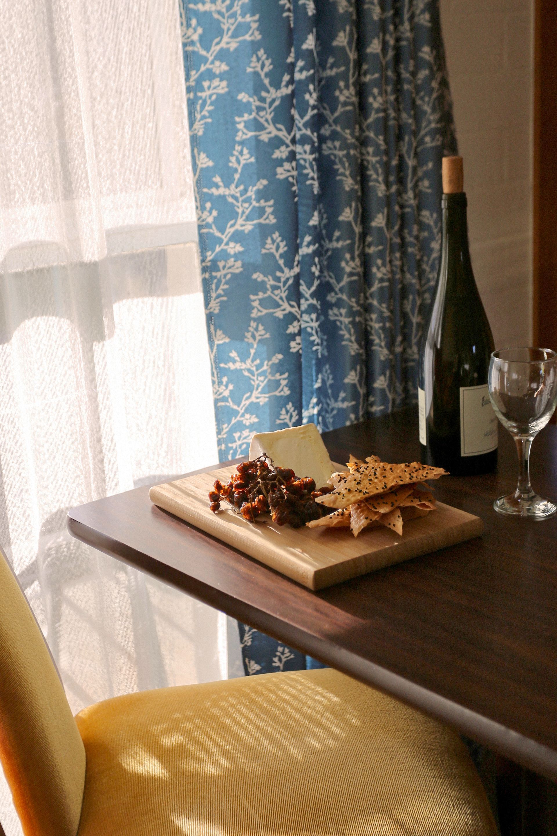 Charcuterie board with cheese, crackers, and jam next to a wine bottle and glass on a small table near a window.