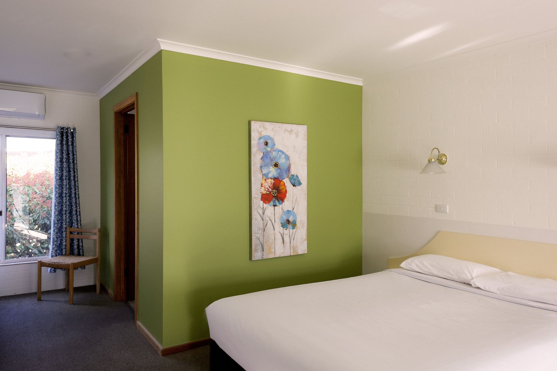 Bedroom with a bed, green wall with art, window with a chair, and a door to a side room.