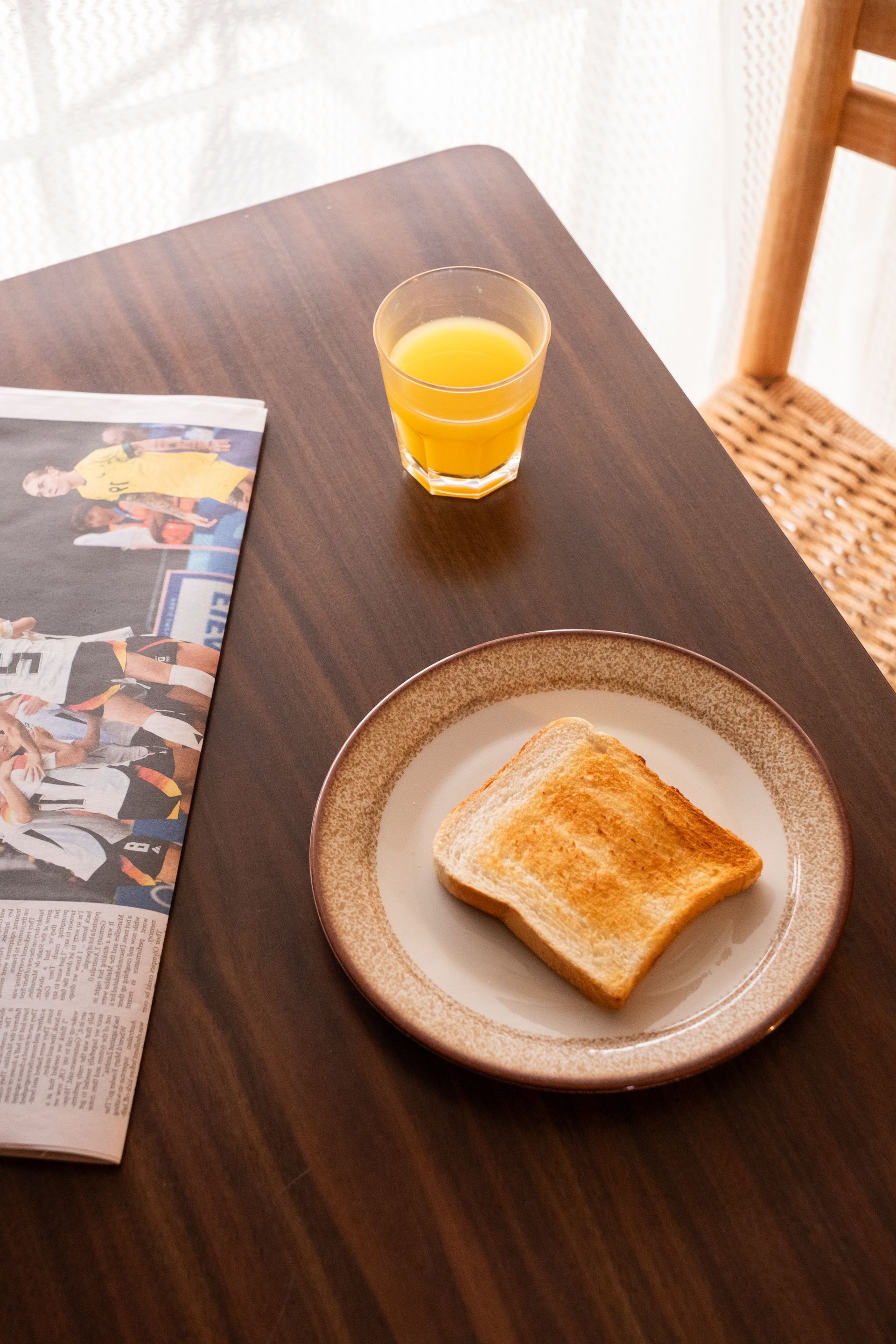 Toast on plate, glass of orange juice, newspaper on a dark wooden table near a window.