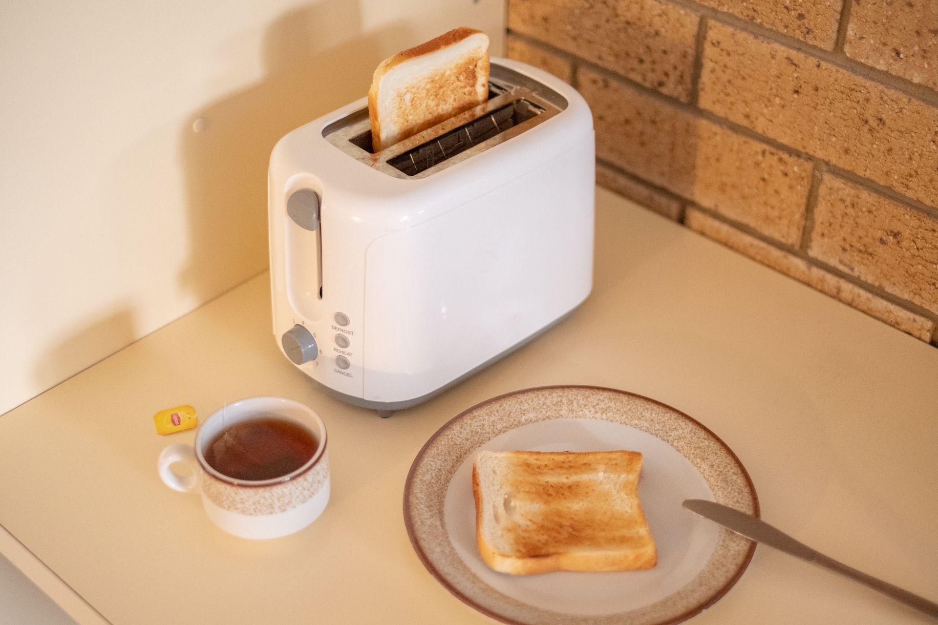 White toaster with toast popping up, alongside a plate of toast and a cup of tea.
