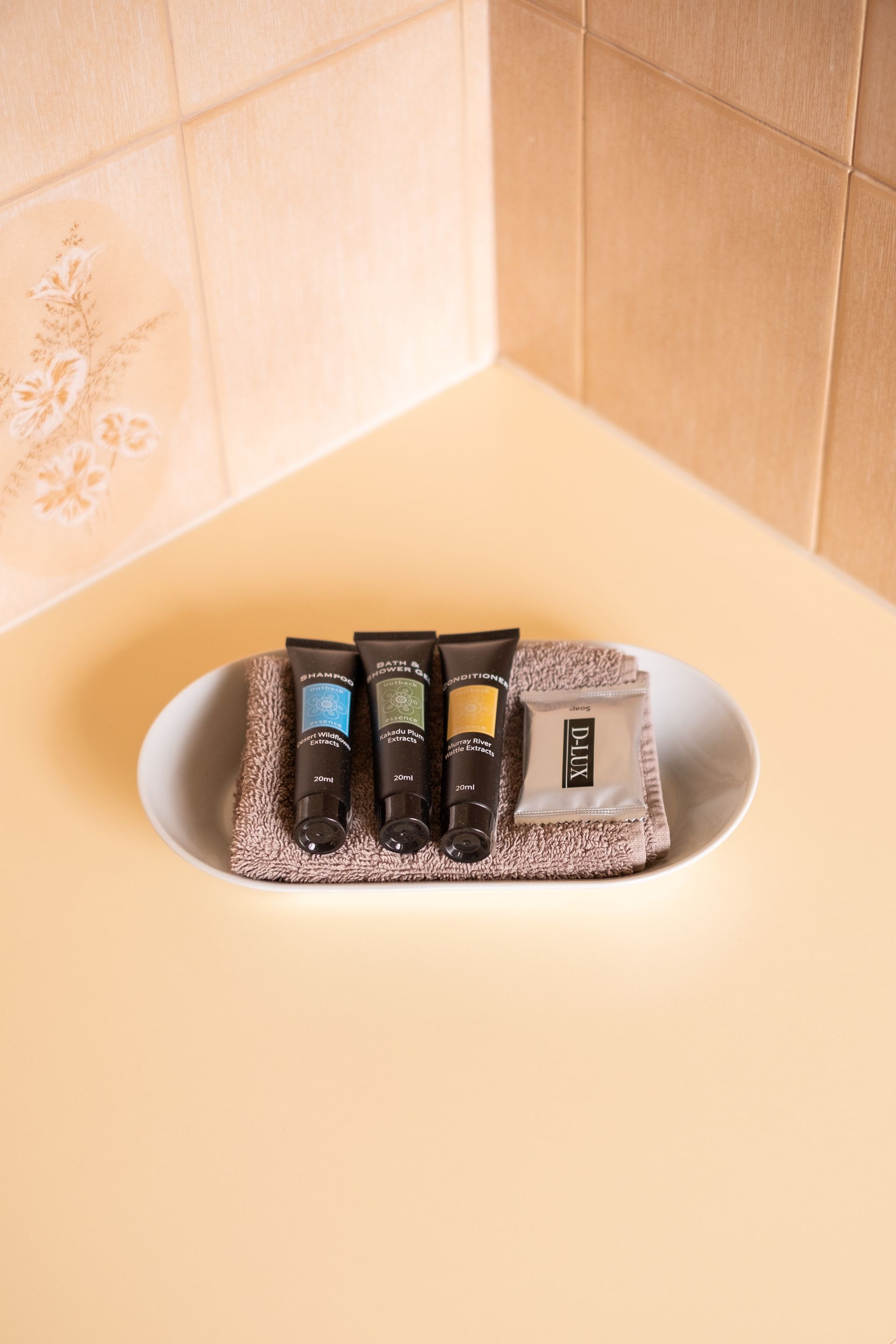 Hotel toiletries in an oval dish: lotions, washcloth, and soap, set on a beige tiled corner.