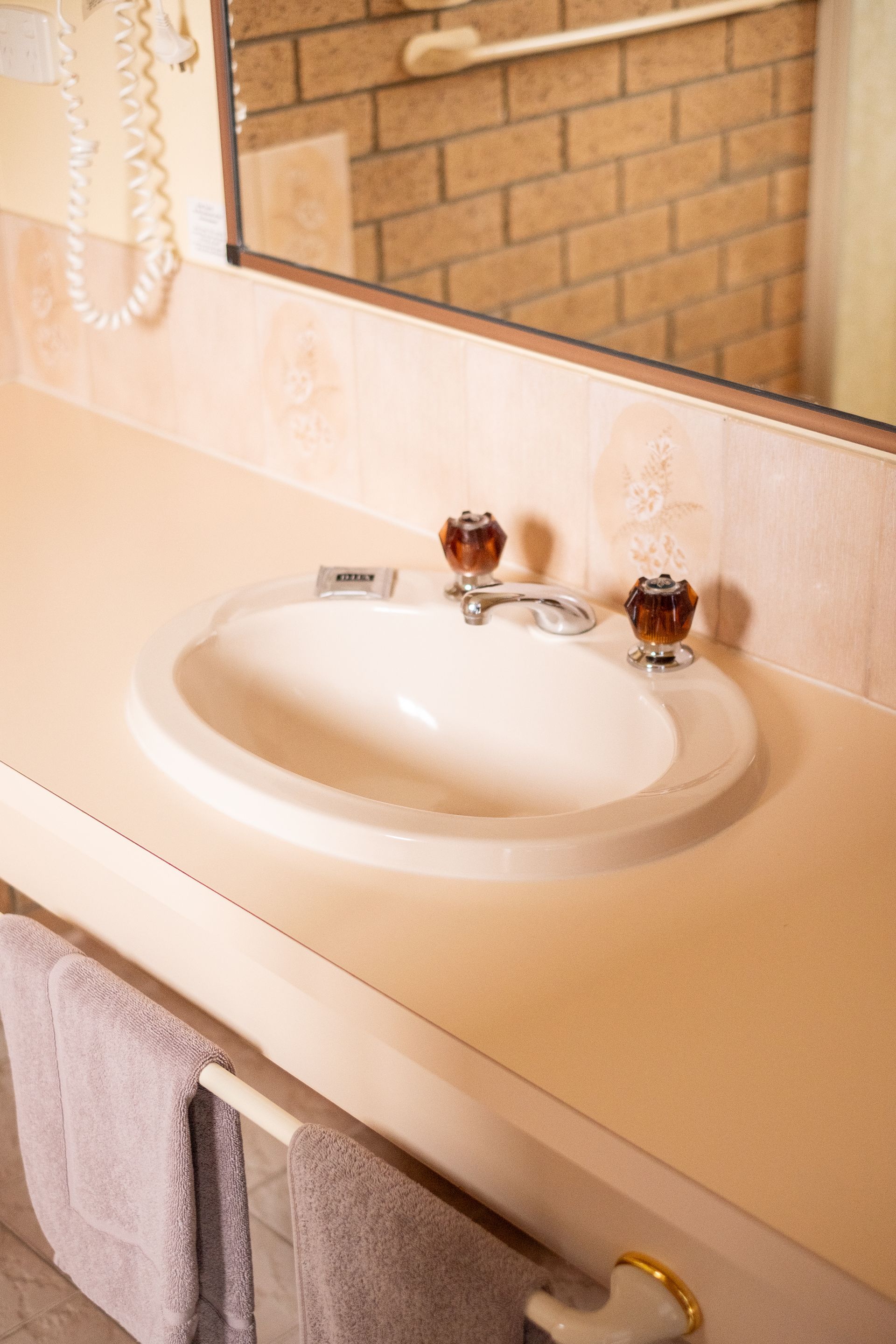 Bathroom sink with beige countertop, brick wall reflected in mirror. Towels on rack.