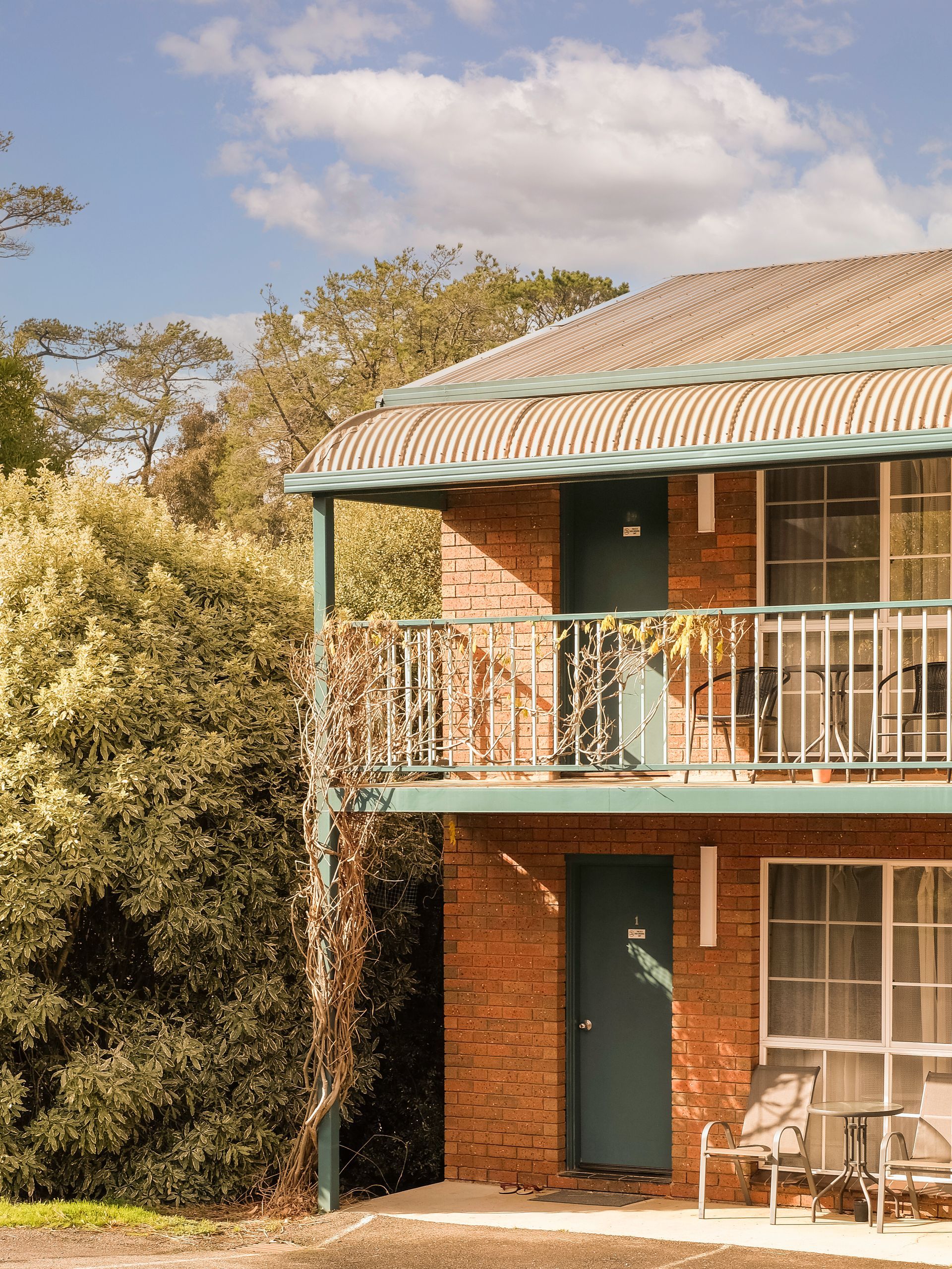 Two-story brick building with teal doors and balcony, set among trees under a cloudy sky.