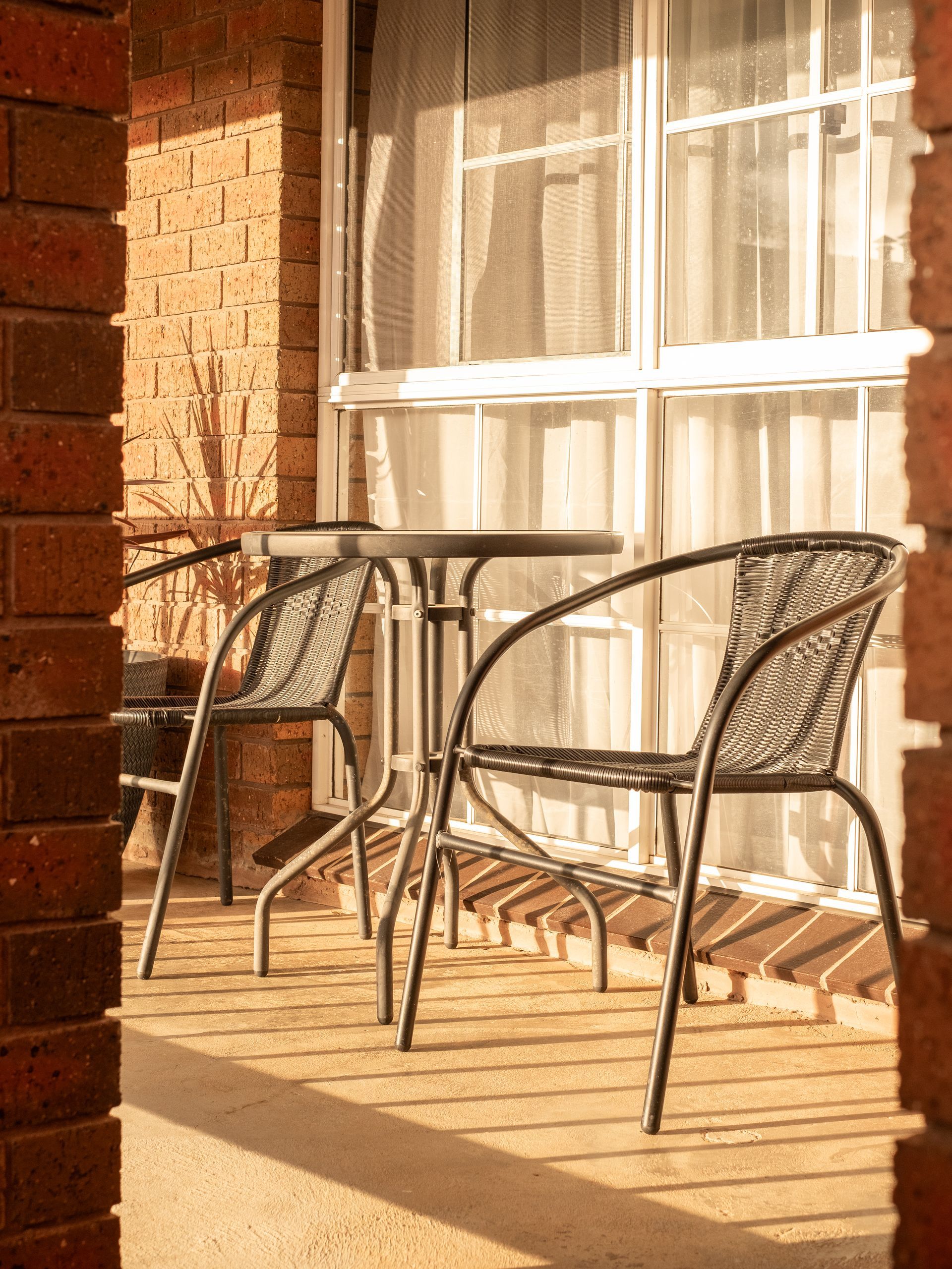 Two black metal chairs and a table on a brick patio in front of a window with sheer curtains.