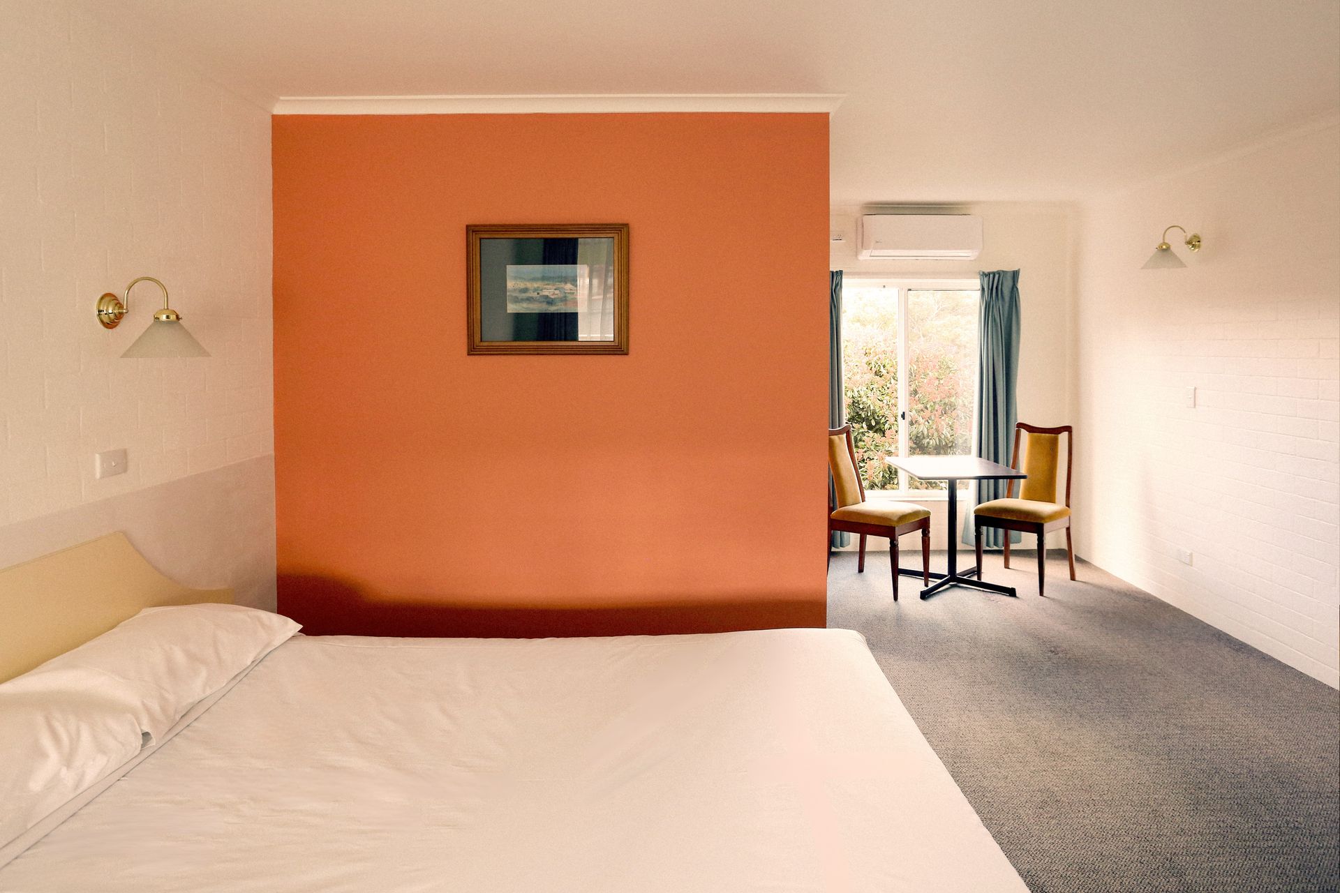 Bedroom with bed, orange accent wall, table and chairs by a window.