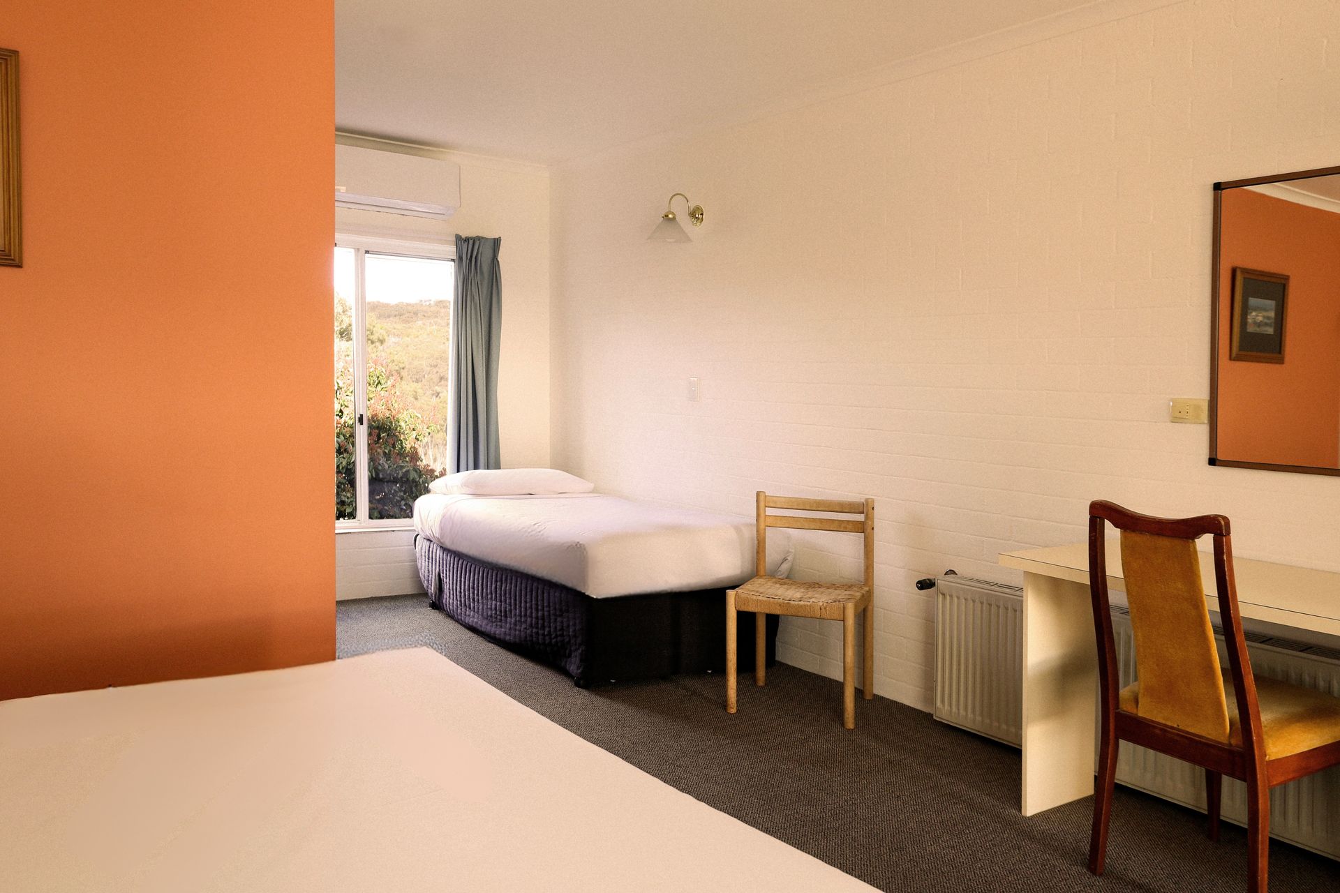 Bedroom interior with beds, desk, chair, and a window with a view of greenery. Orange and white walls.