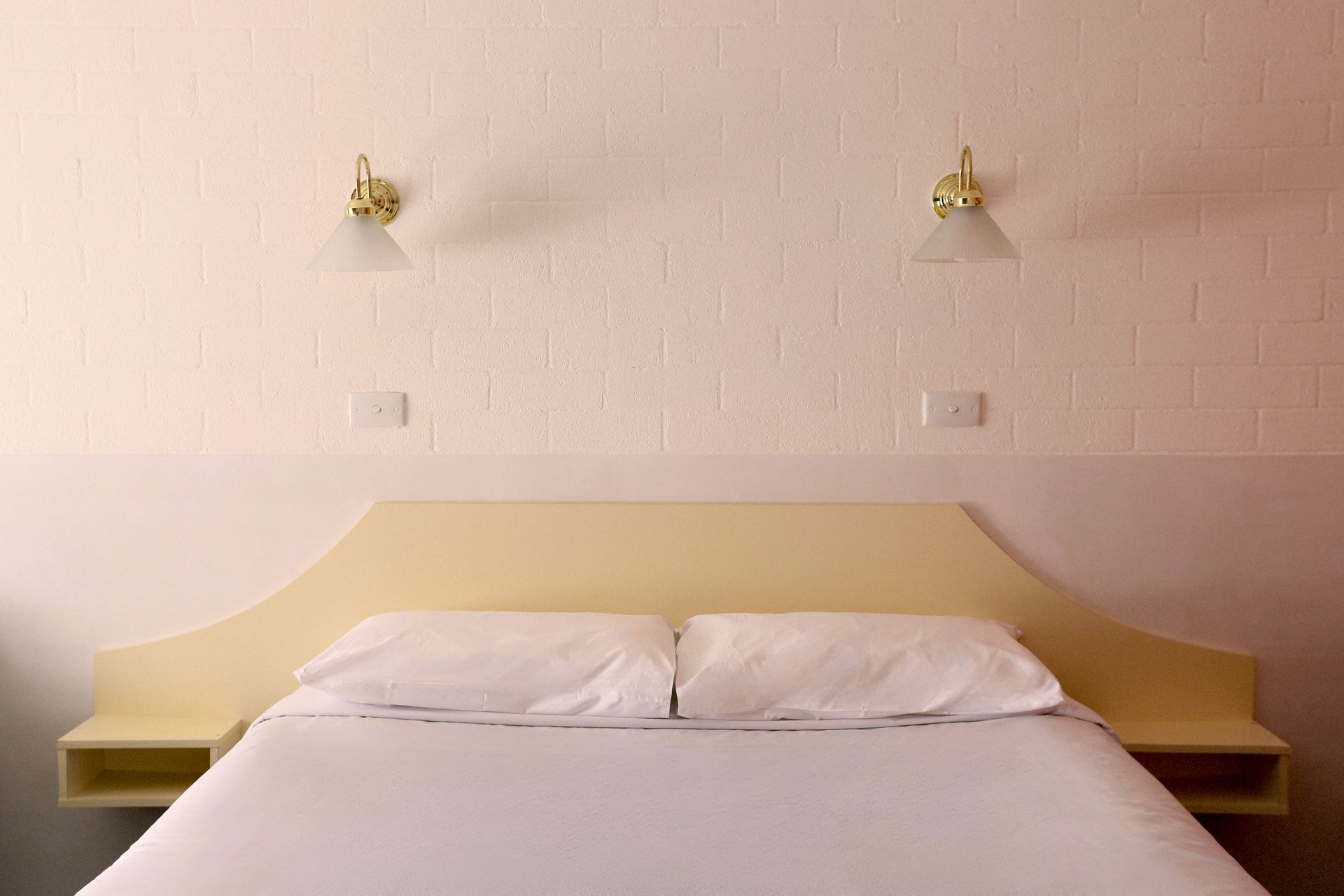Bedroom with bed, white bedding, beige headboard, wall lights, and white walls.