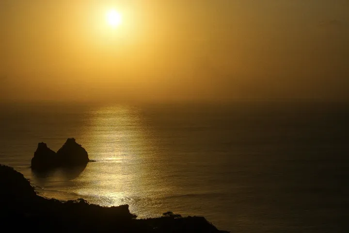 O sol está se pondo sobre o oceano com duas rochas em primeiro plano.