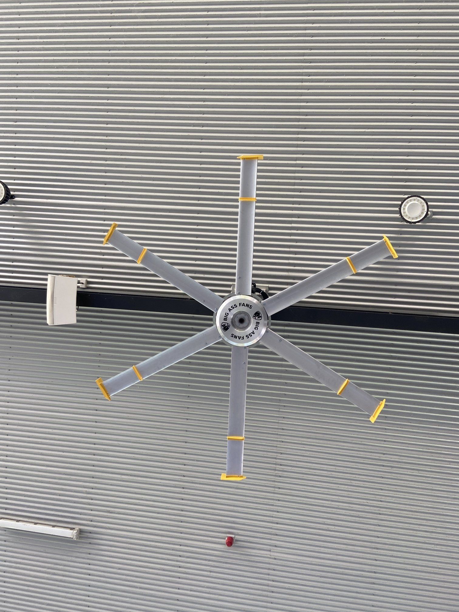 Ceiling-mounted gray fan with yellow-tipped blades against a corrugated metal ceiling.