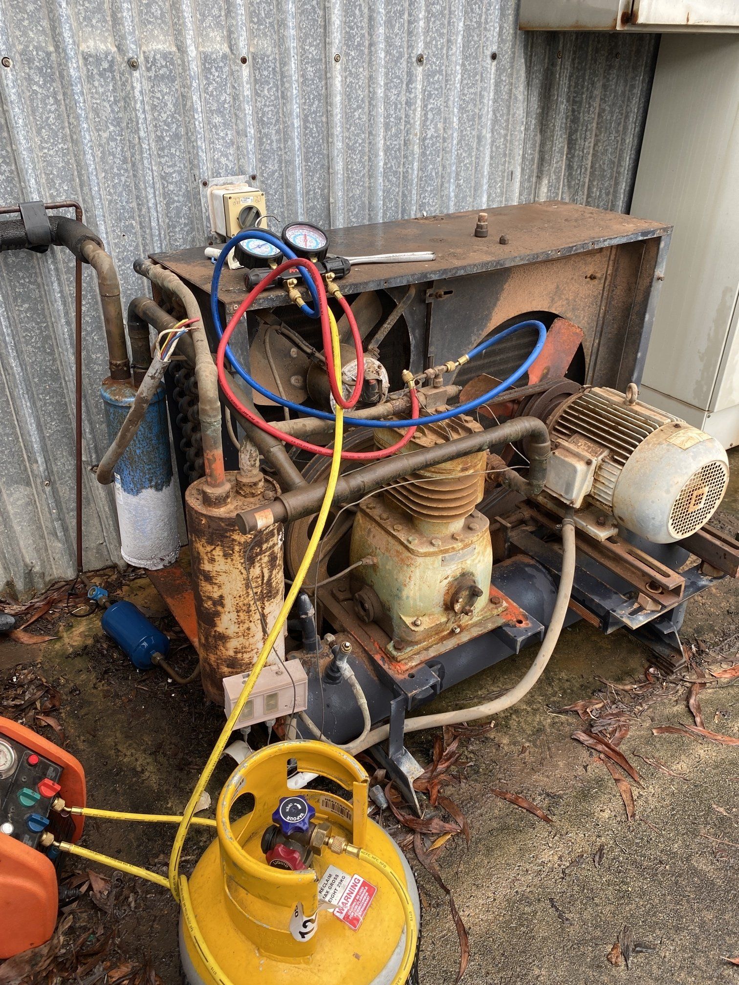 Old industrial air compressor with tangled hoses and yellow tank against a corrugated metal wall