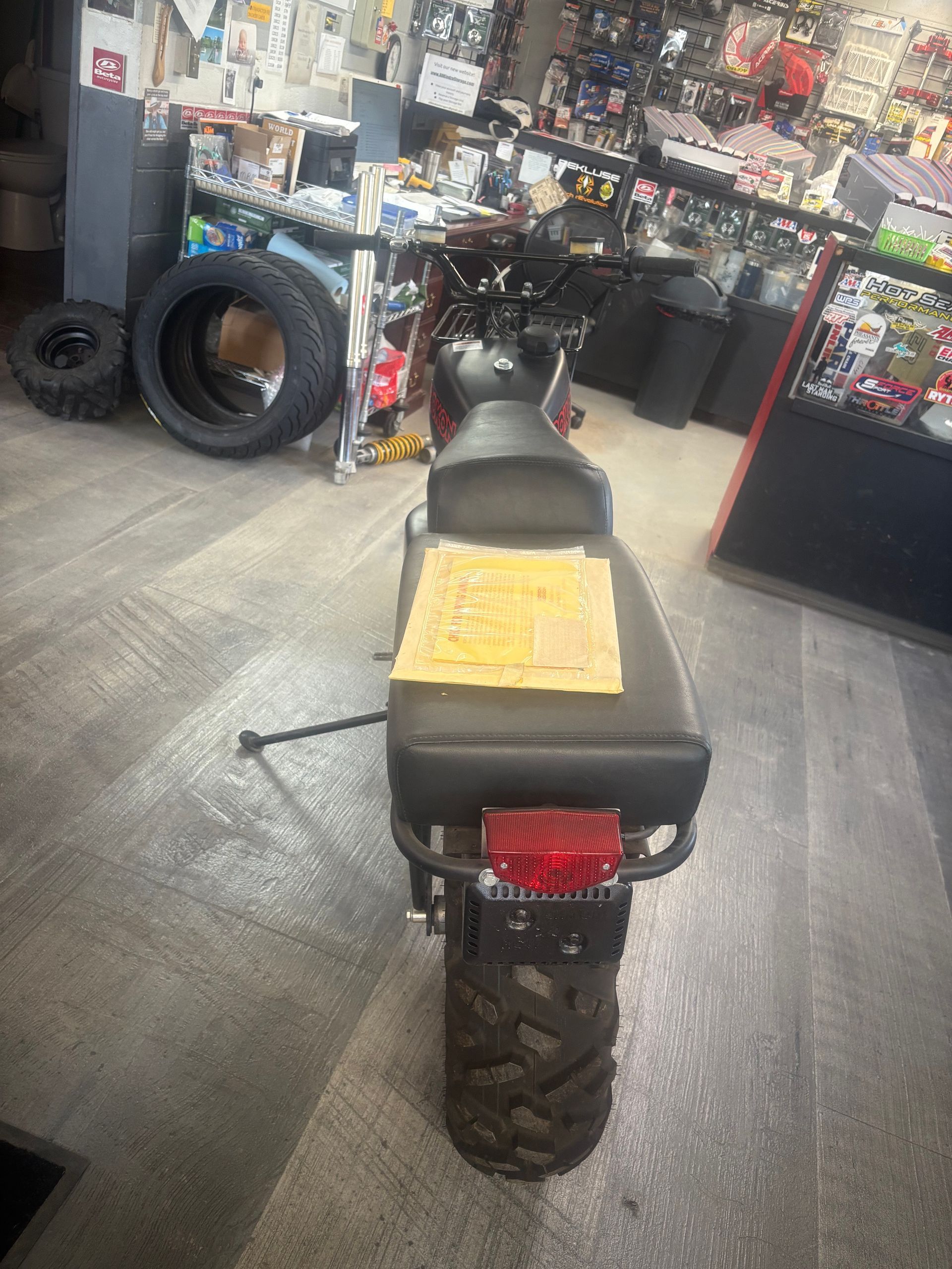 Black dirt bike in a shop, viewed from the rear, with a yellow seat cover and motorcycle tires nearby.