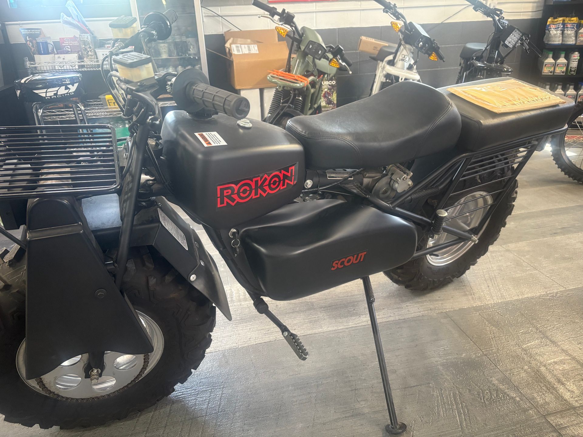 Black REKON electric scooter with seat and rear cargo rack displayed indoors.