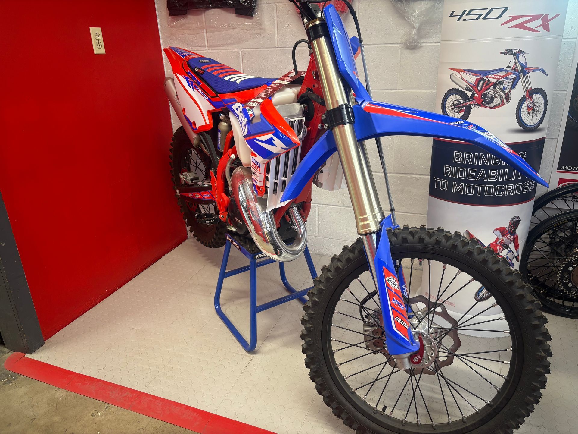 Blue and red dirt bike on a display stand in a showroom, with a red wall and floor accent.