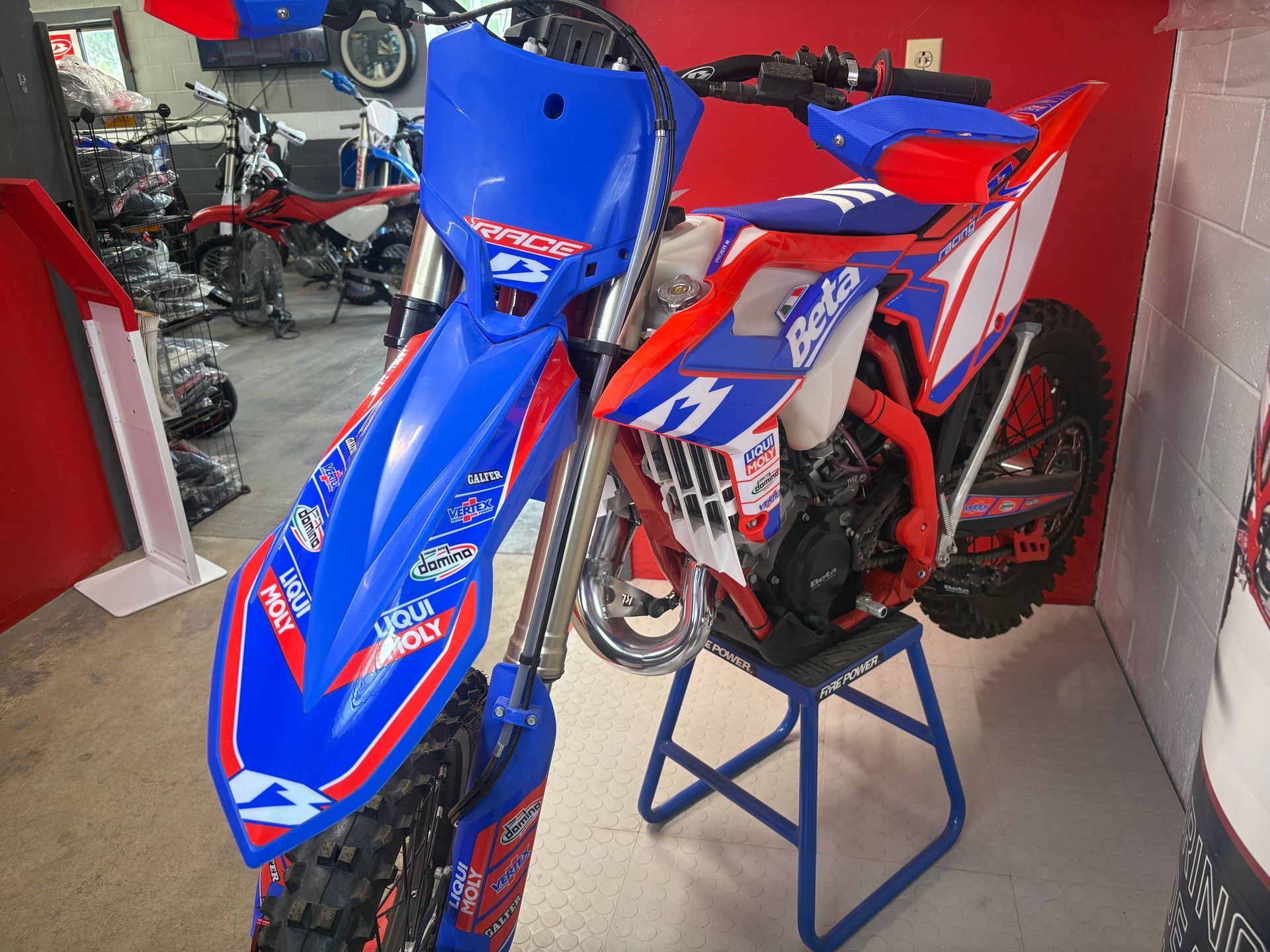 Blue and red dirt bike on a stand in a garage, with another bike in the background