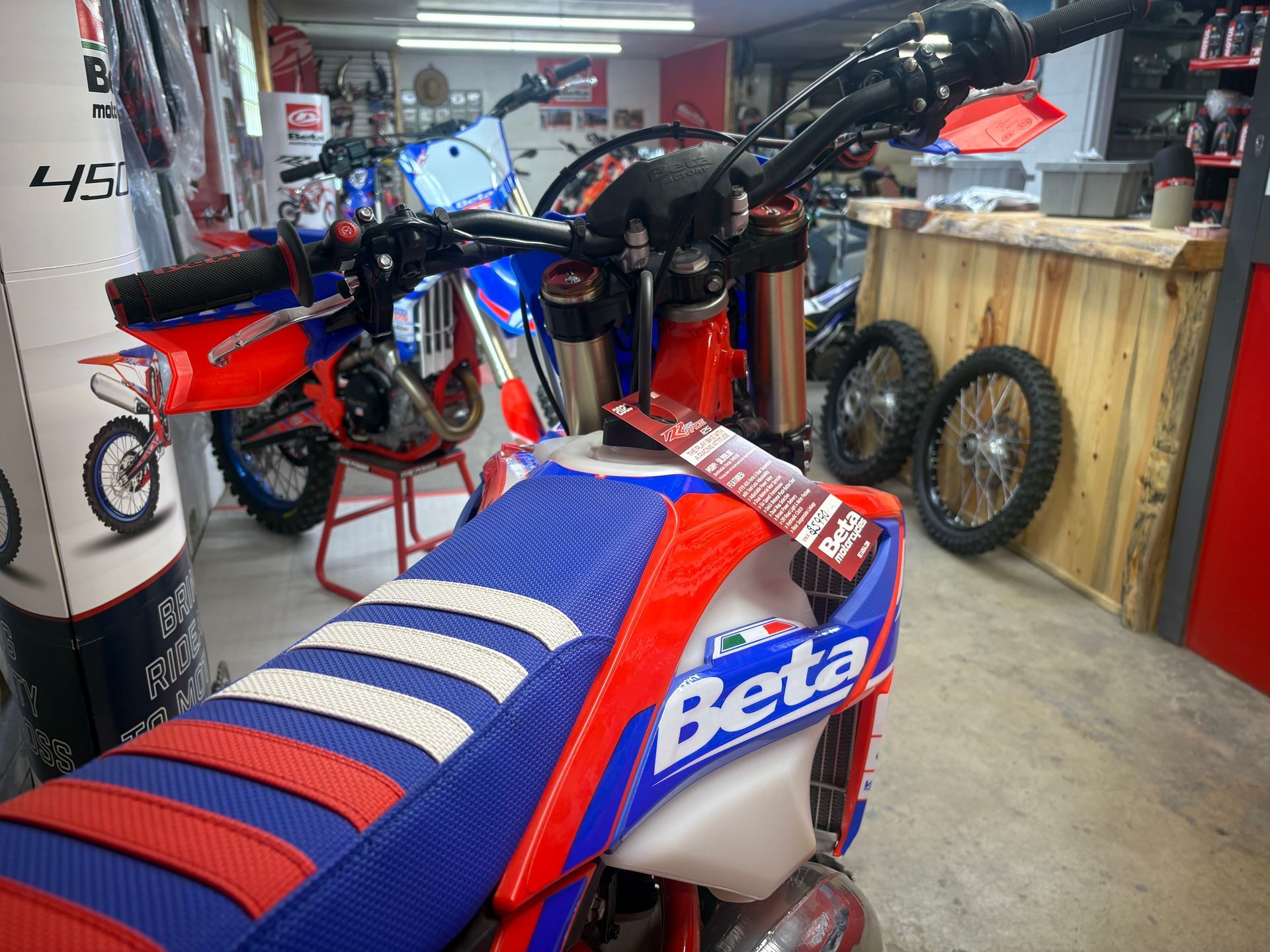 Blue and red Beta dirt bike in a workshop, with a second bike and spare tires in the background