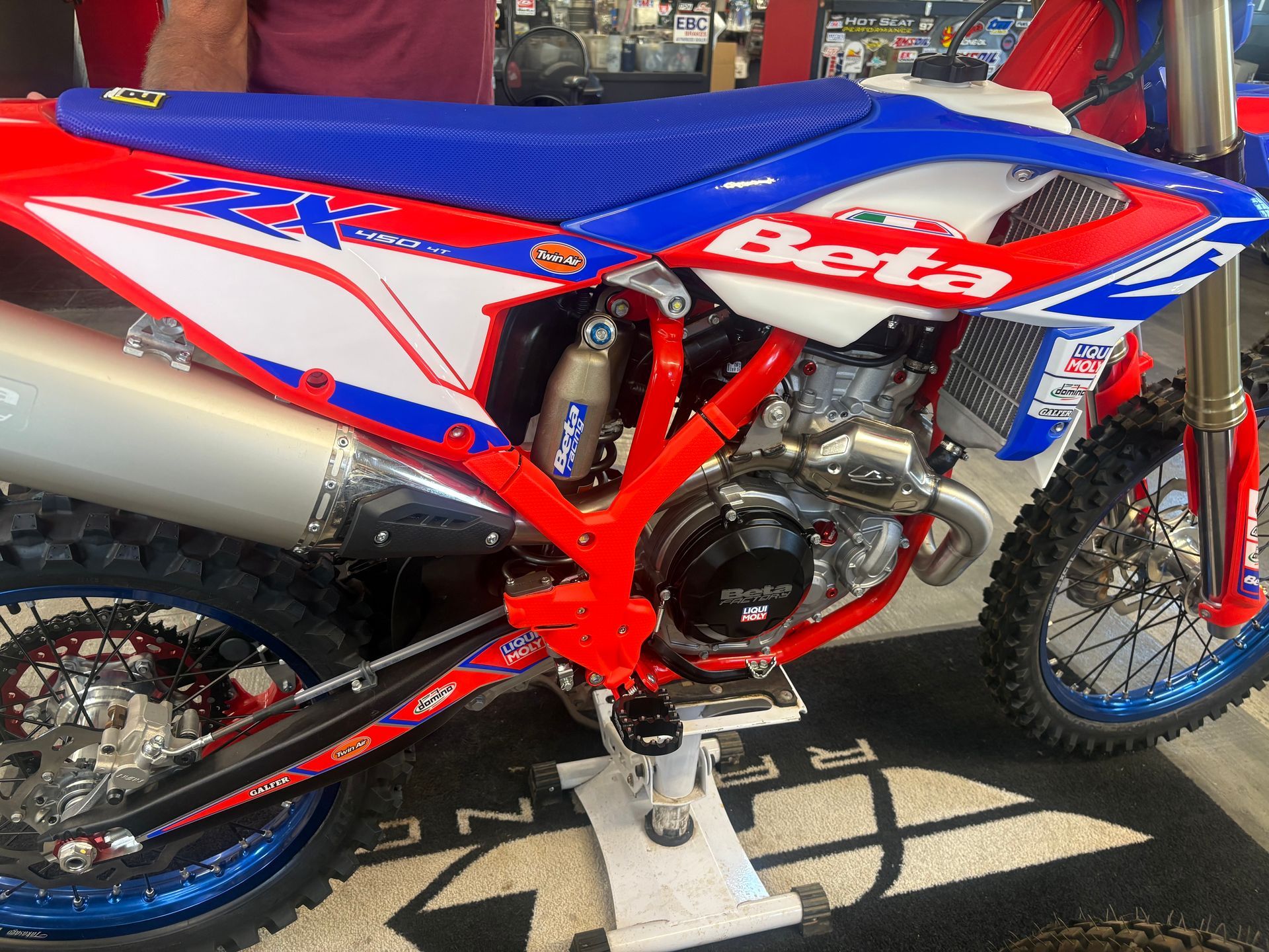 Red, white, and blue Beta dirt bike on a stand, side view in bright sunlight.