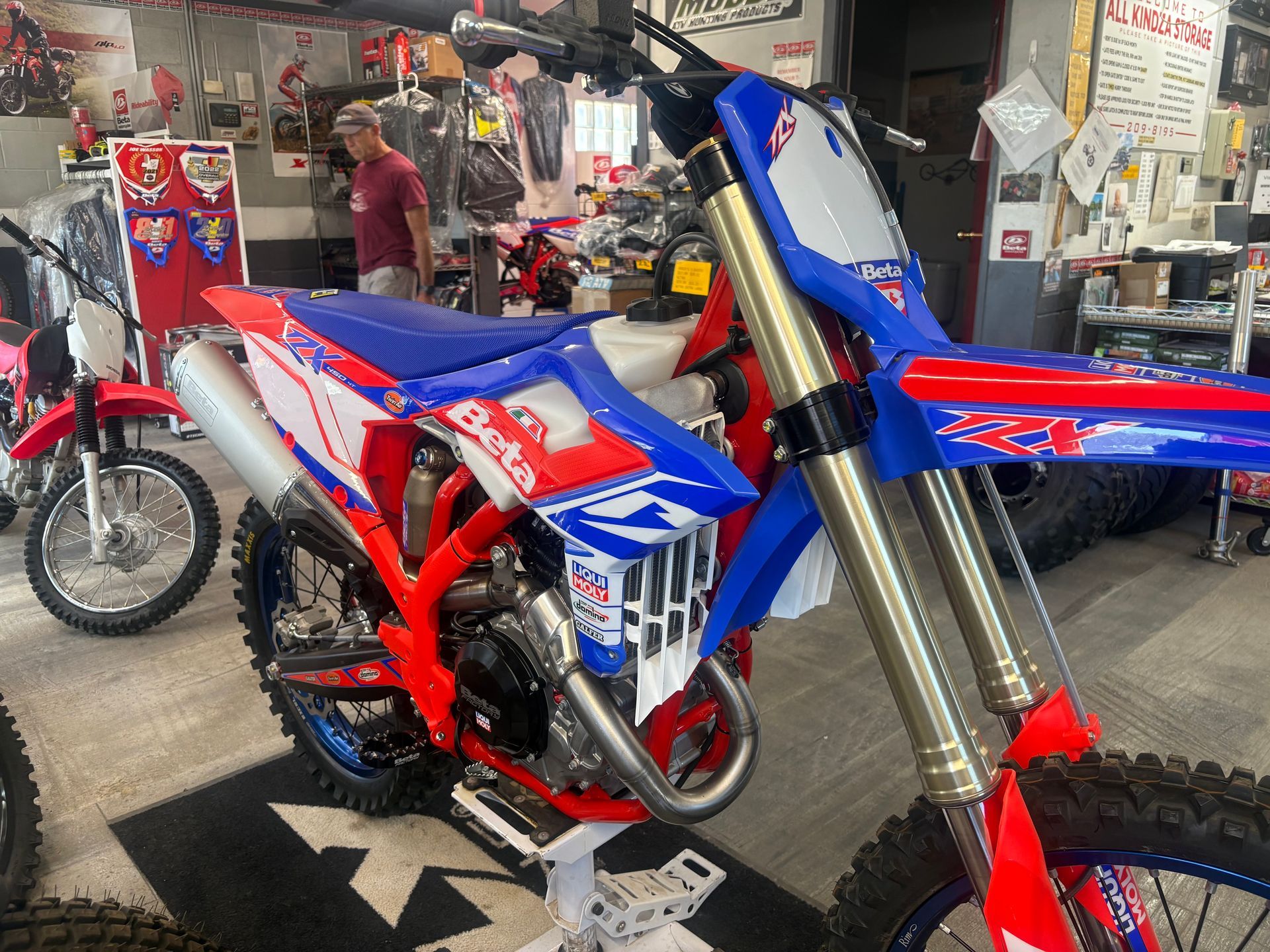 Blue and red dirt bike in a workshop, front view with gold forks and a red stand
