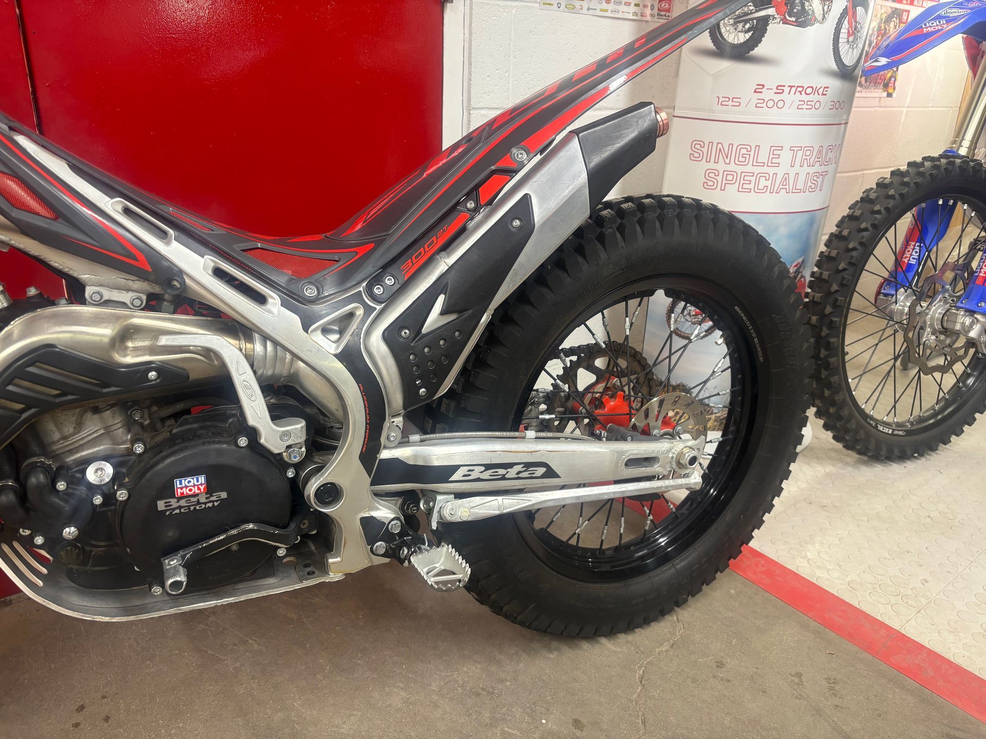 Red and black dirt bike parked indoors, close-up of rear wheel and engine area.